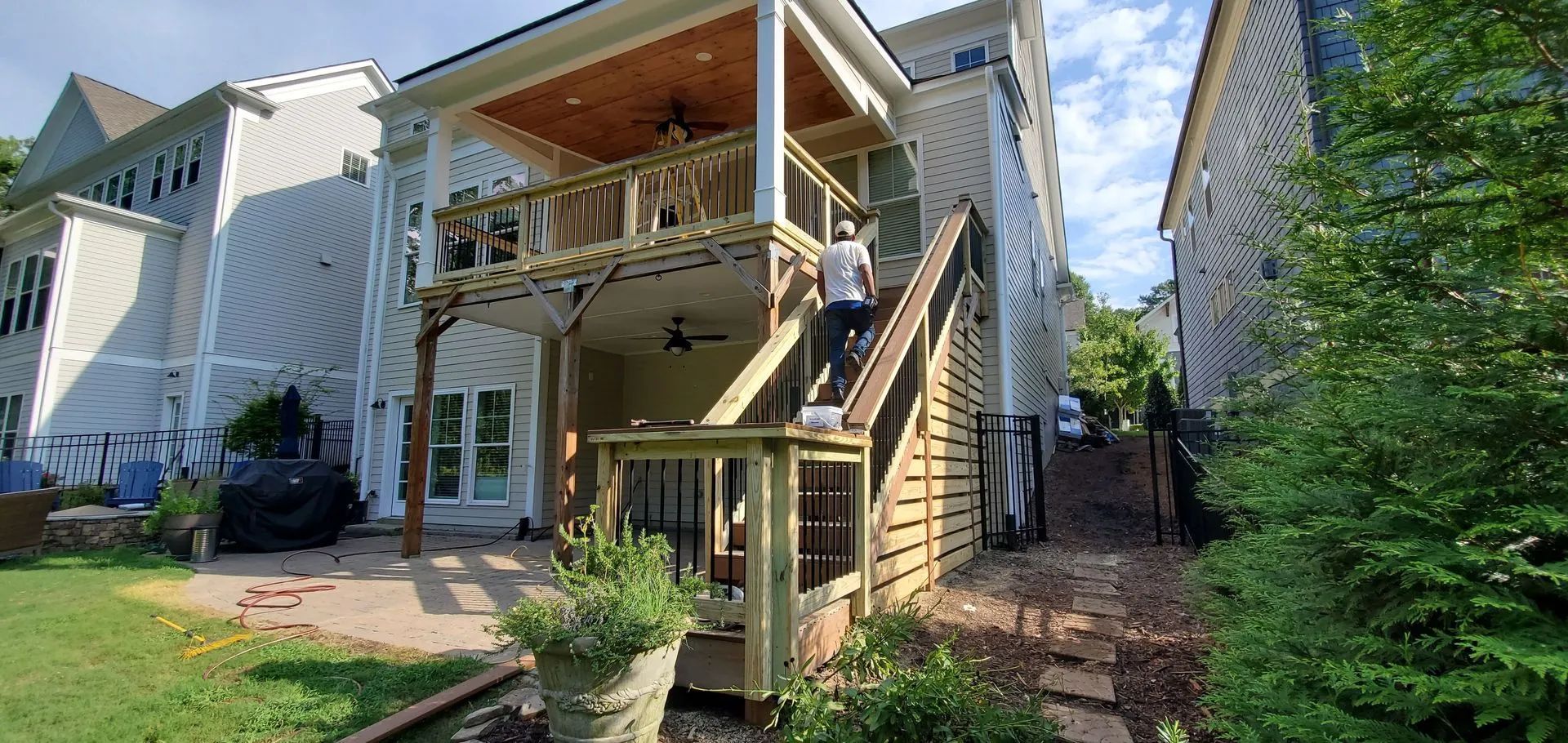 Person climbing wooden stairs to a two-story deck of a house. Green grass and trees surround it.