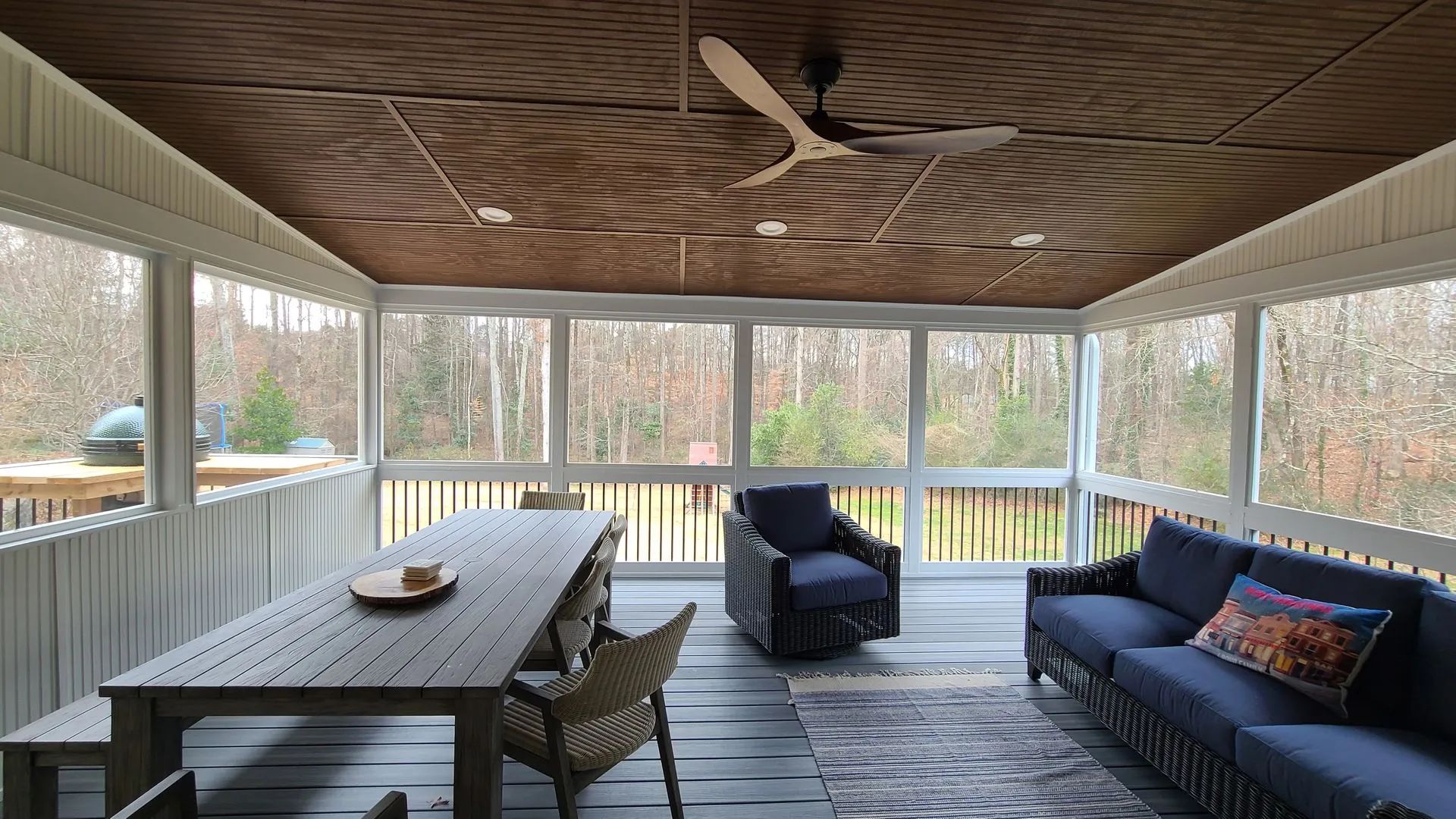 Screened-in porch with blue furniture, wooden table, and fan, overlooking a wooded area.