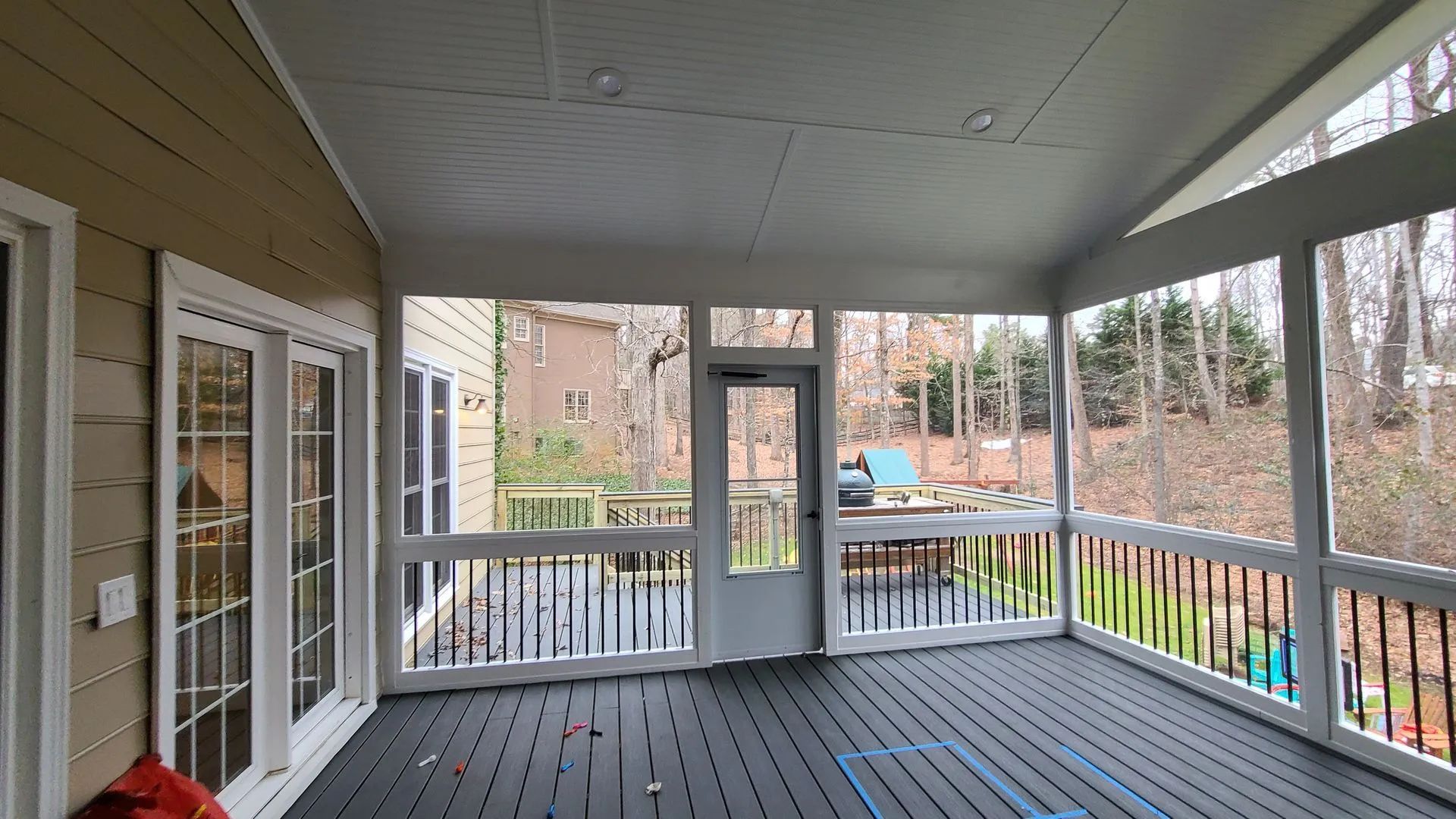 Screened-in porch with black railing, gray deck, and door to backyard. White trim, tan siding, trees in background.