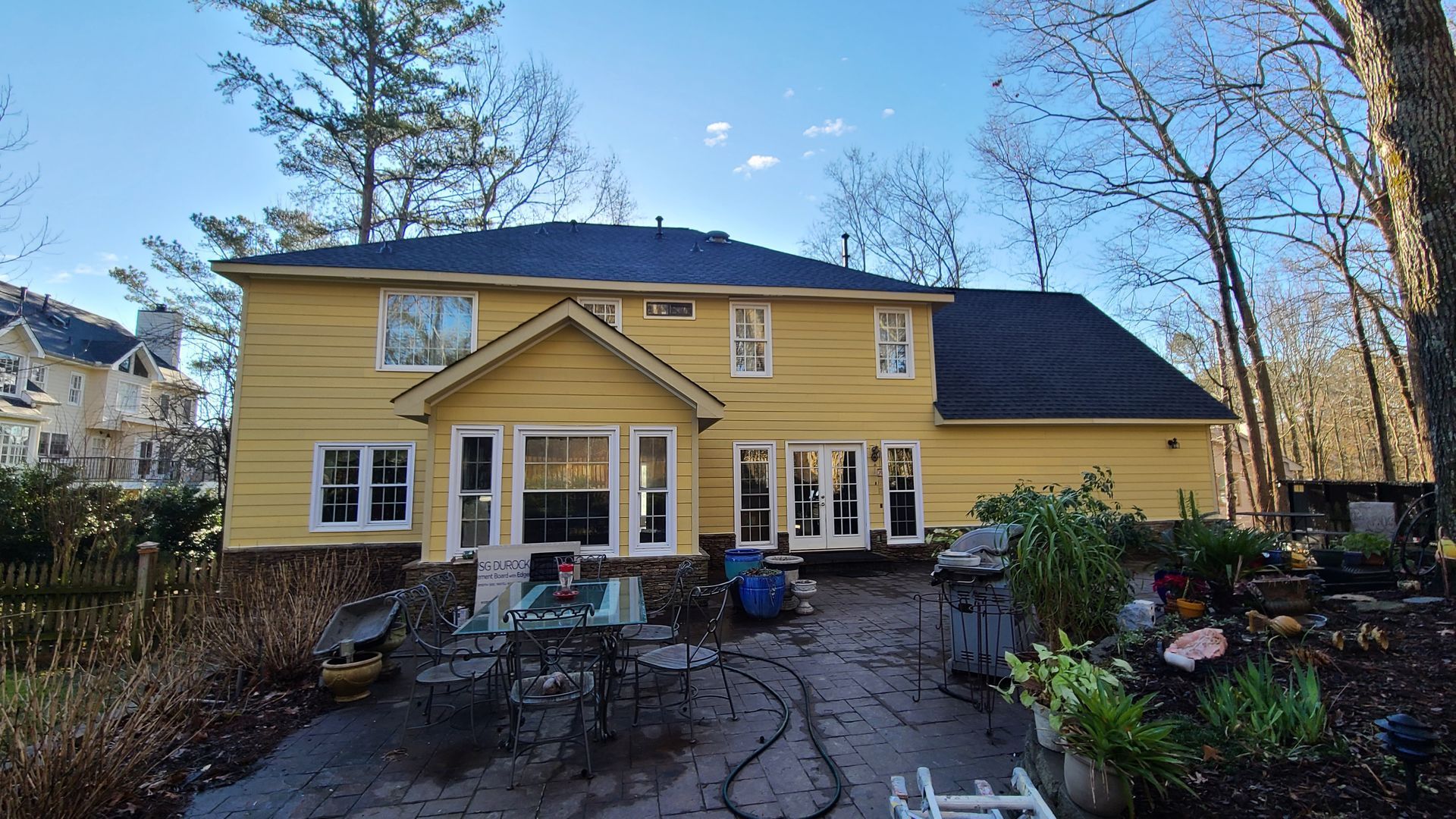 Yellow house with dark roof, patio furniture, and surrounding trees against a blue sky.