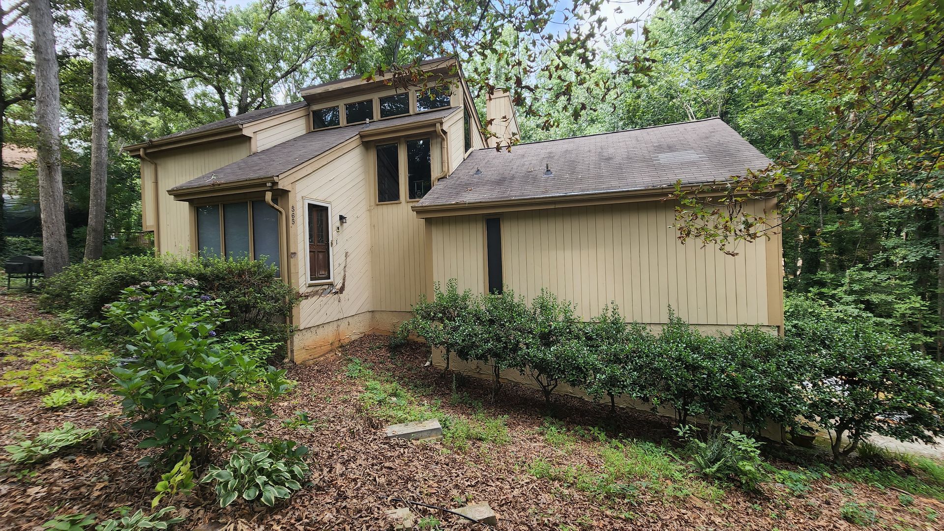 Beige house with angled roof, surrounded by trees and bushes, on a slight hill.