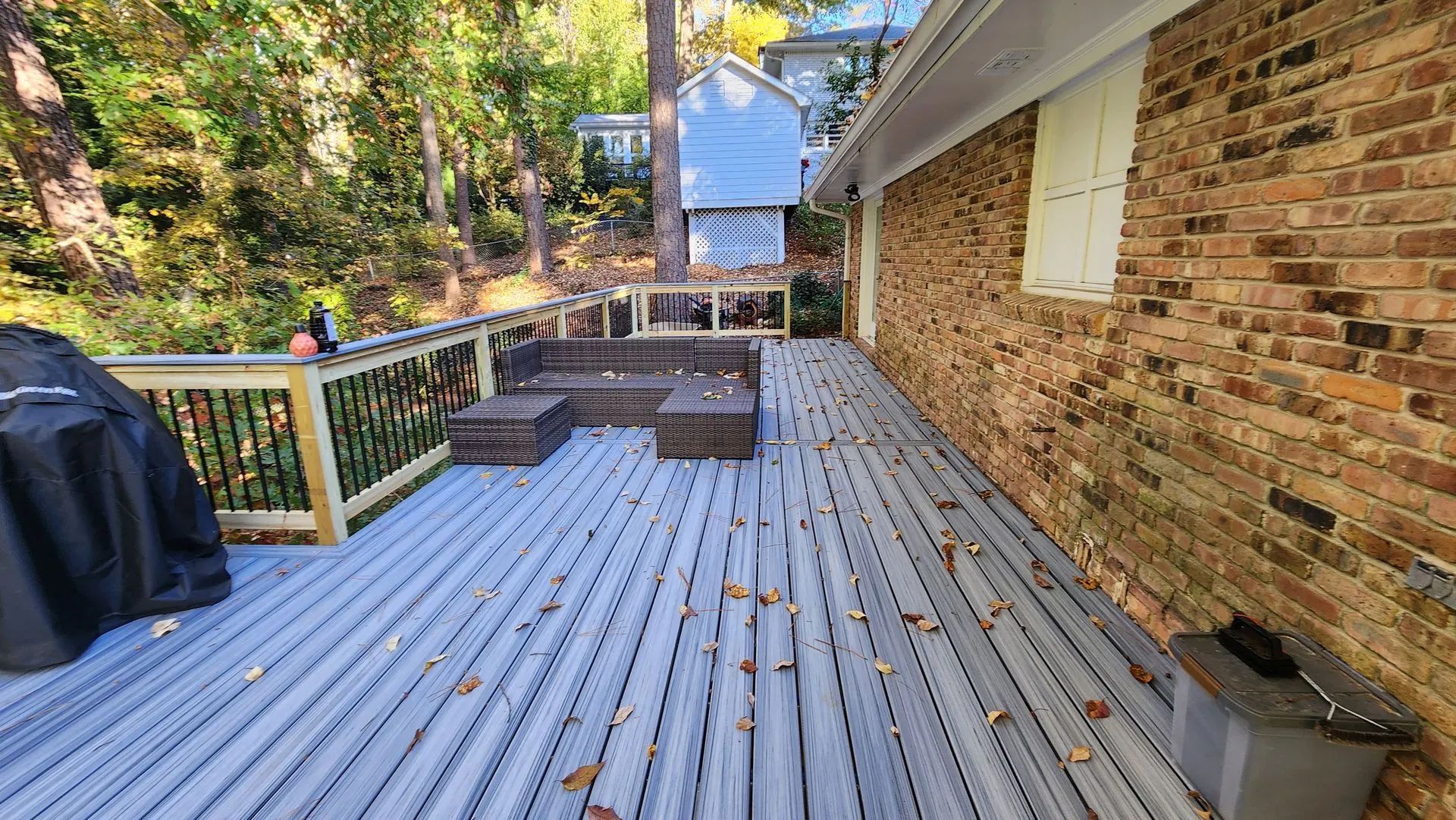 A grey deck with a brick wall, wooden bench, and grill in a yard surrounded by trees.