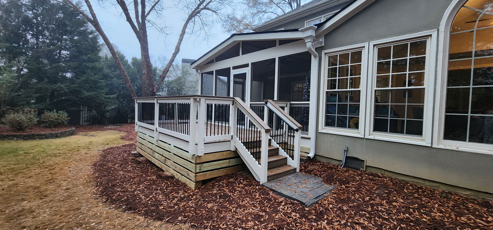 Screened porch with stairs, next to a house with windows, surrounded by mulch and grass.