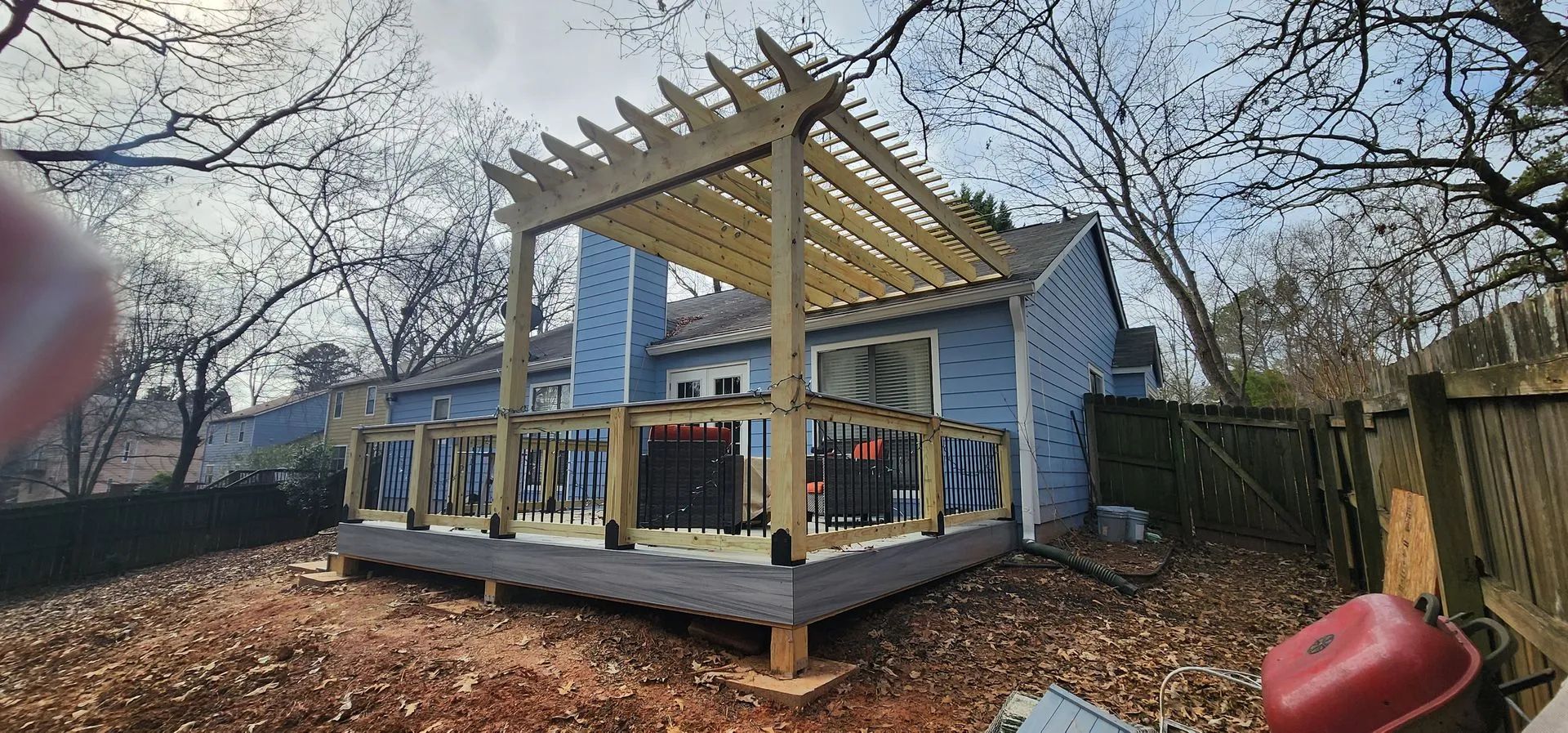 Newly built wooden deck with pergola, blue house in background, surrounded by bare trees, dirt ground.