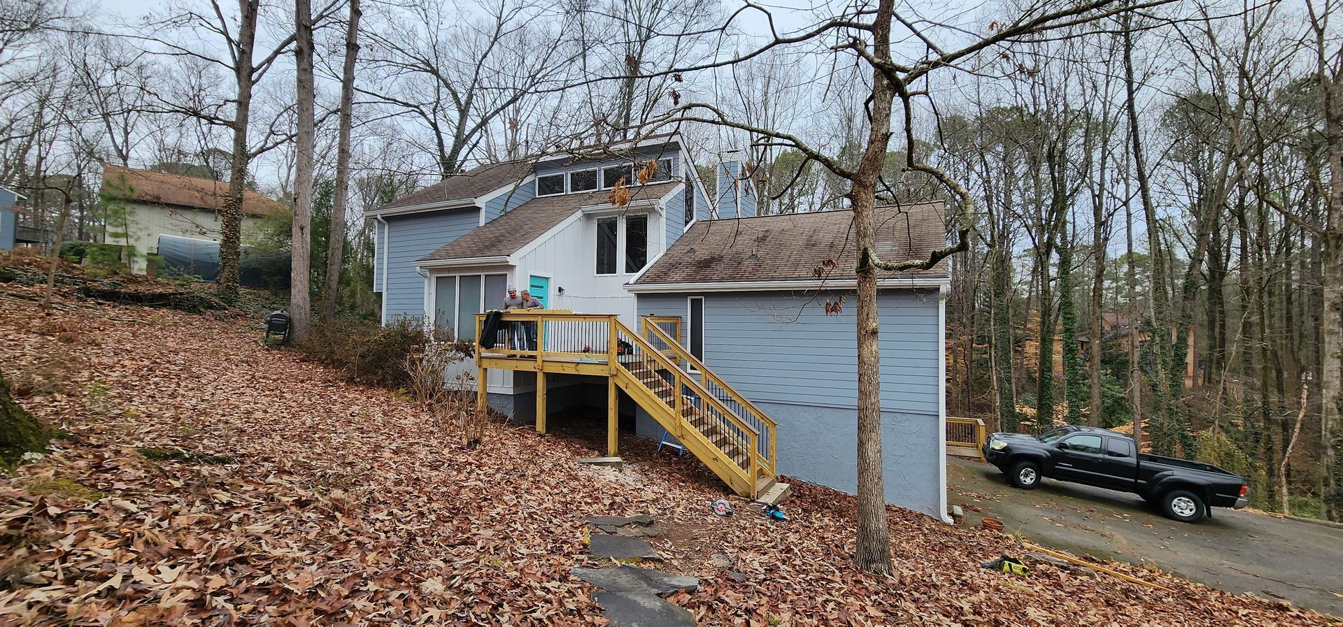 A house with a deck, garage, and black truck on a driveway, surrounded by trees and brown leaves.