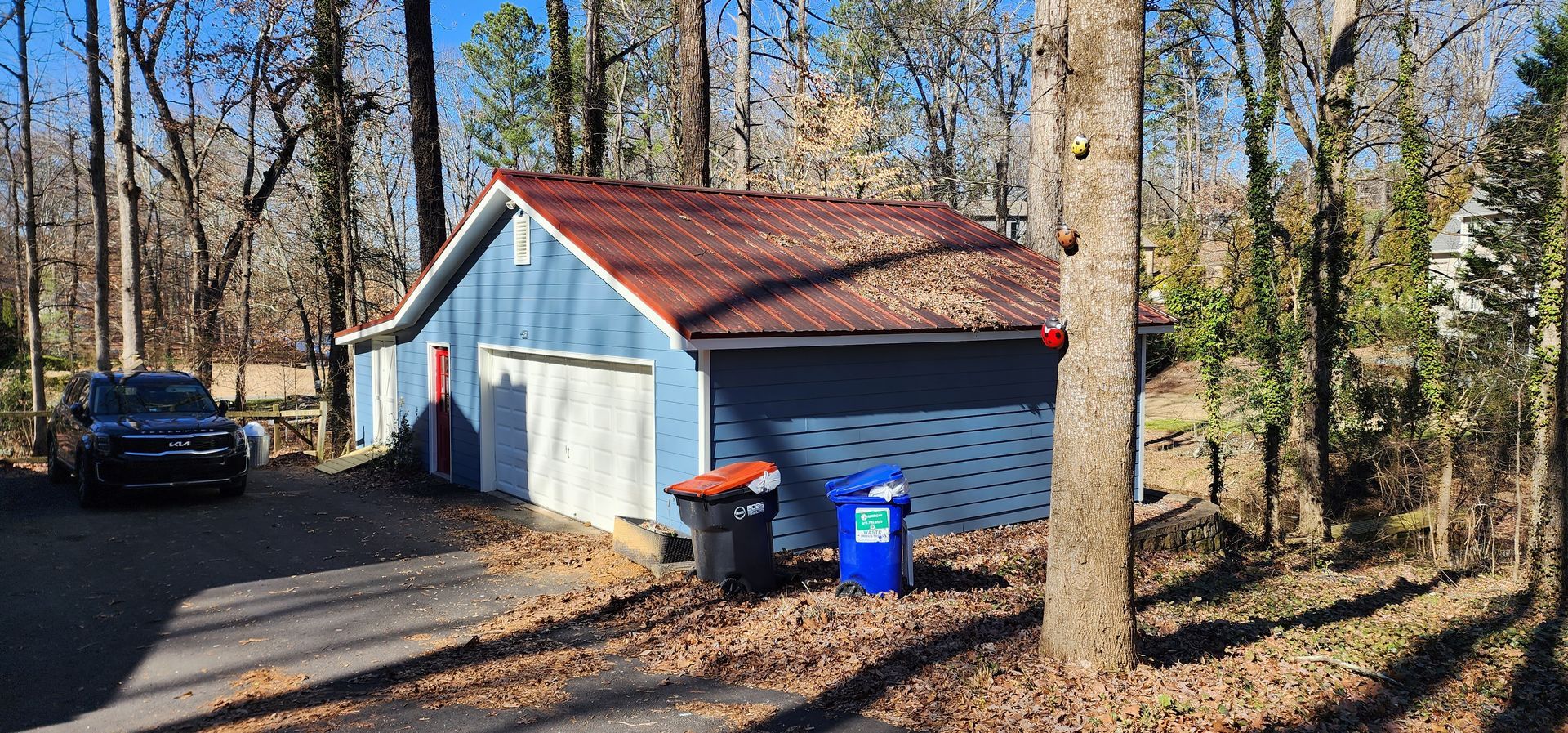 Blue garage with red roof, surrounded by trees, and a dark blue car in the driveway.