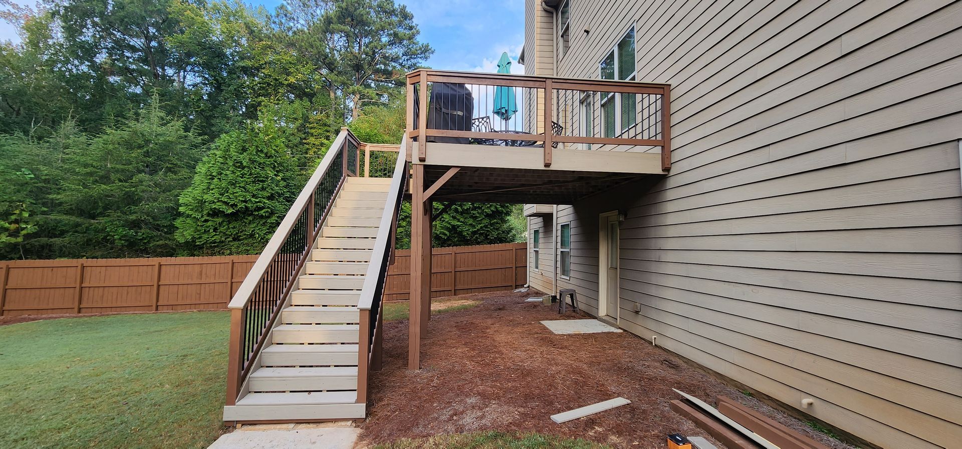 A wooden deck with stairs leading down to a yard with grass, a brown fence, and a building.