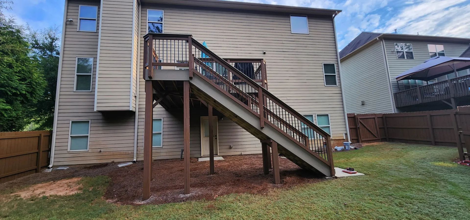 Back of a two-story house with a wooden deck and stairs. Brown siding and a fenced backyard with grass.