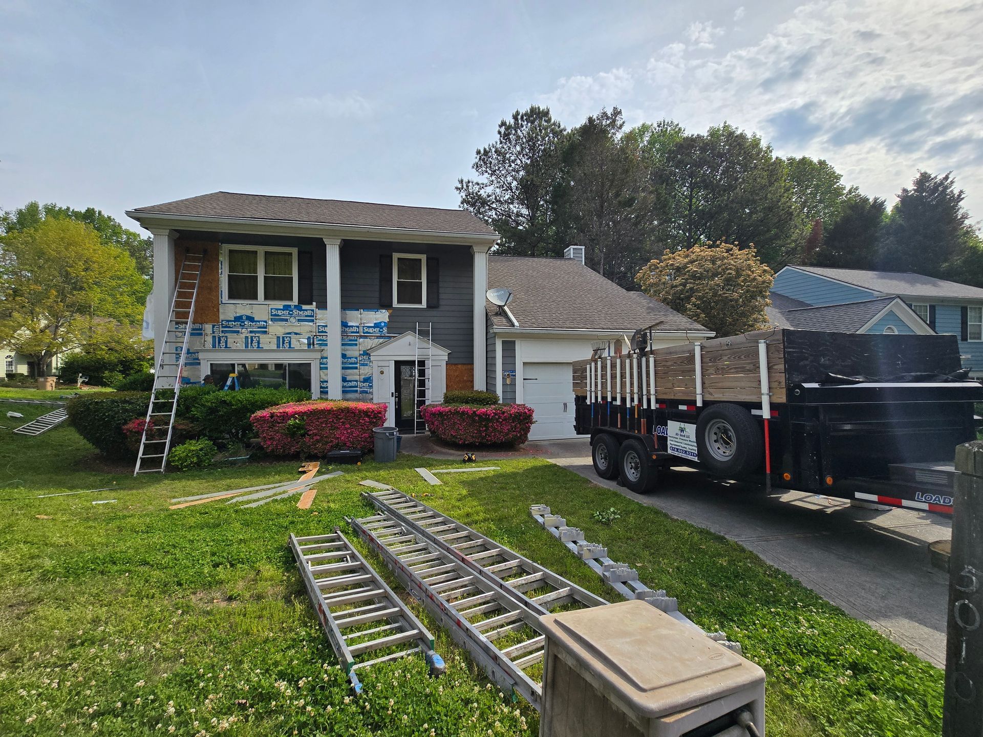 House undergoing exterior renovation, with ladders, trailer, and landscaping.