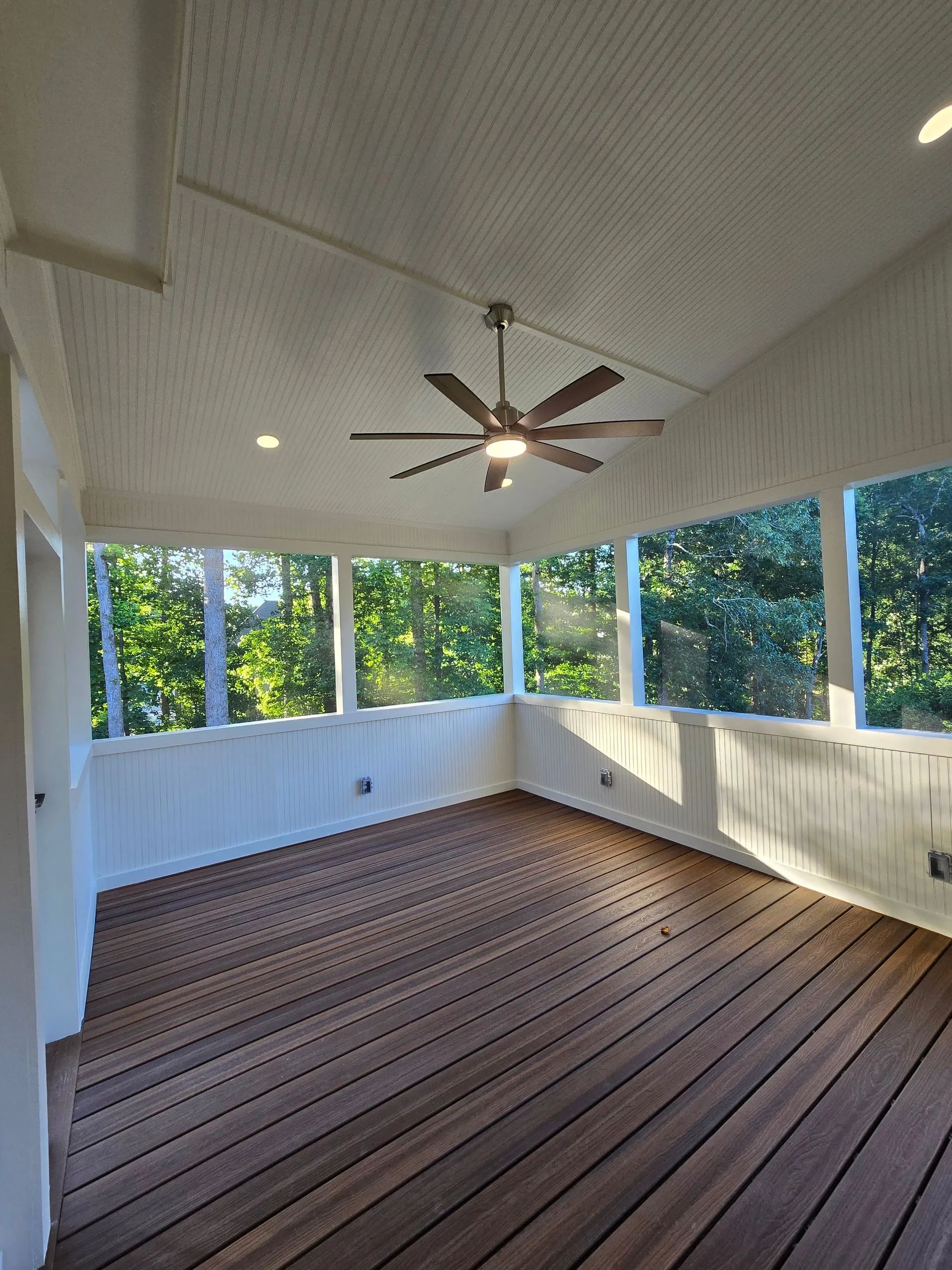 Screened-in porch with wood floor, ceiling fan, and windows overlooking trees.