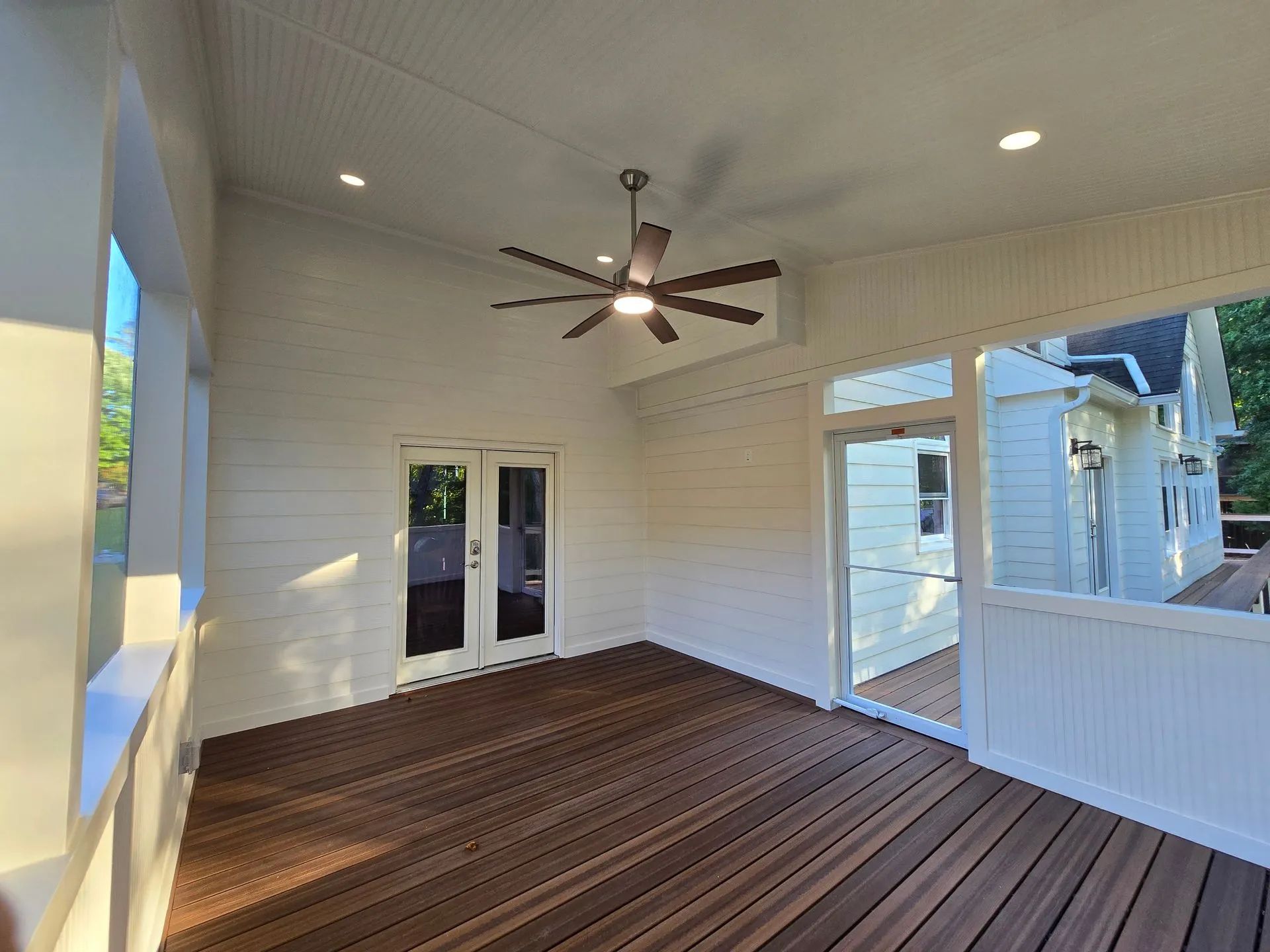 Enclosed porch with dark wood floor, white walls, and ceiling fan. Sunlight streams in from the left.