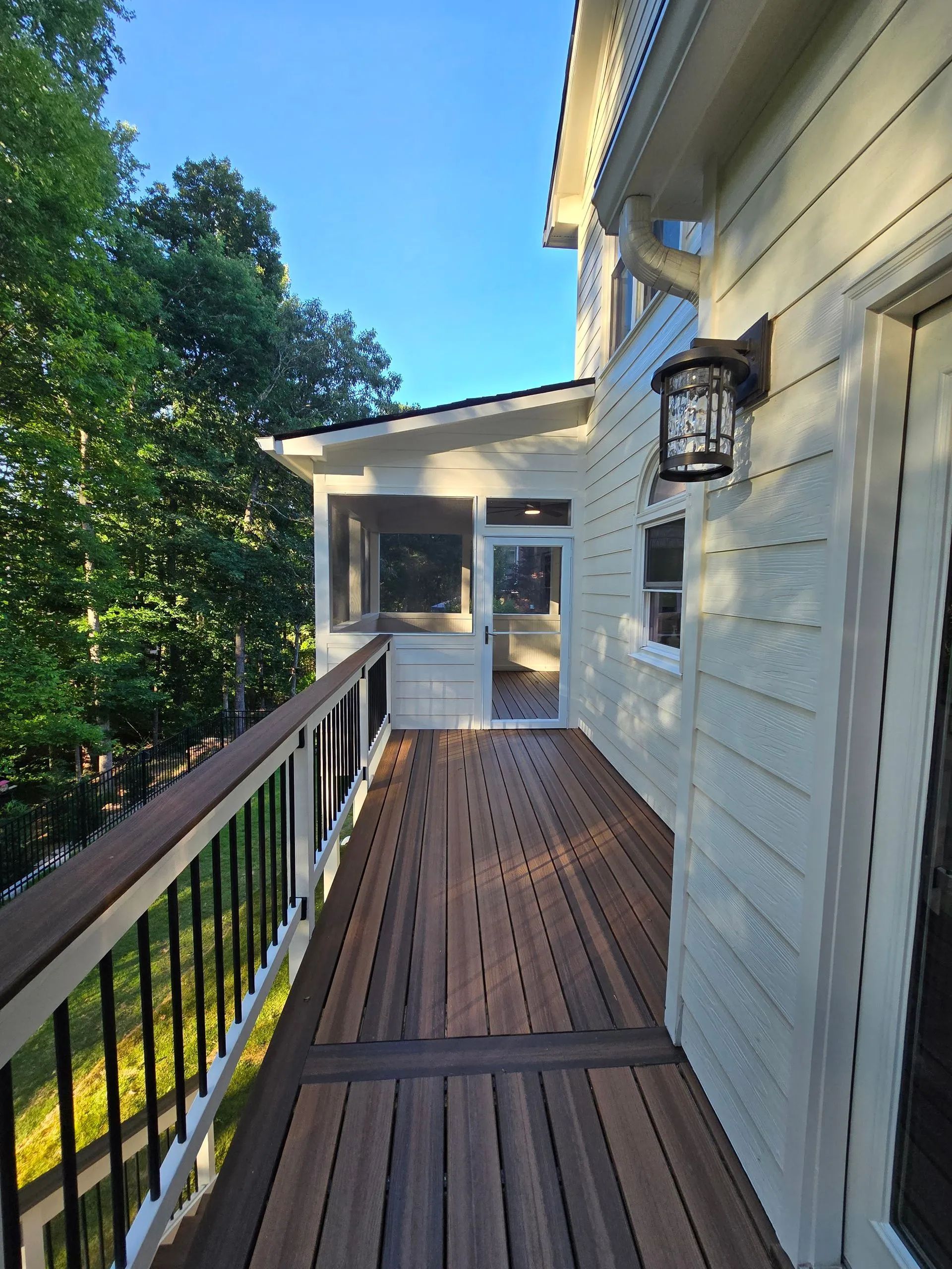Wooden deck with black railing leads to screened porch next to a light-colored house; trees in the background.