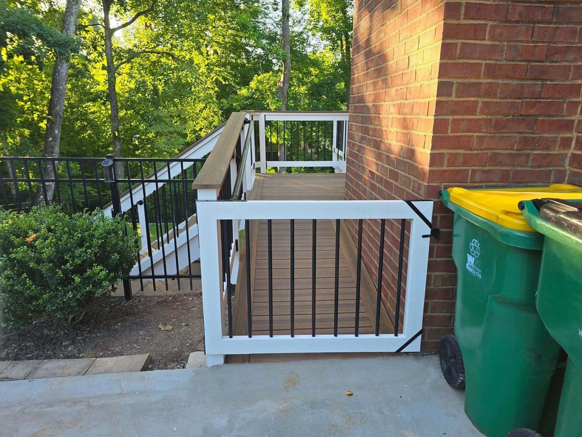 Deck with white railing, black gate, and stairs next to brick wall. Green trash bins.