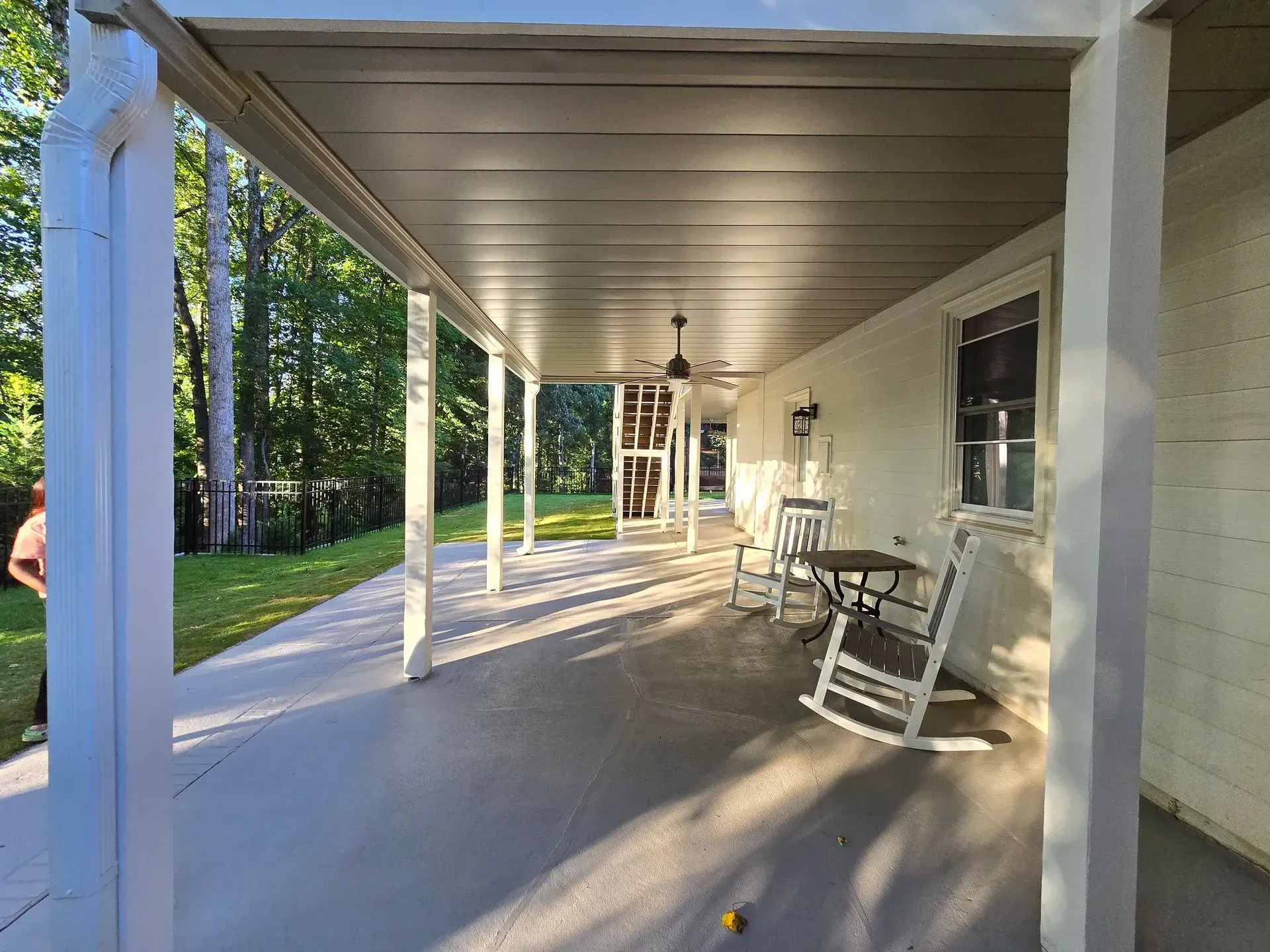 A long, covered porch with a concrete floor, white pillars, and rocking chairs; trees in the background.