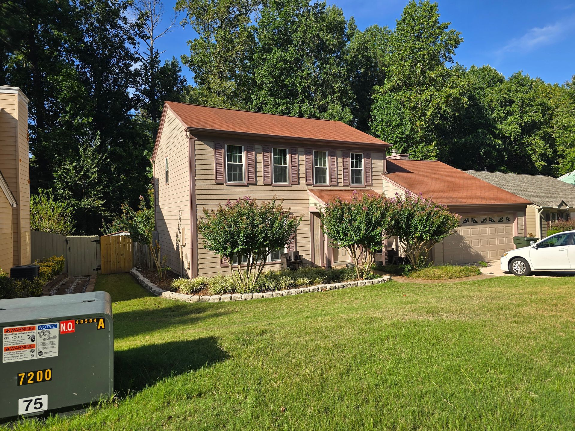 Two-story beige house with brown roof and shutters, set on a green lawn with trees in the background.