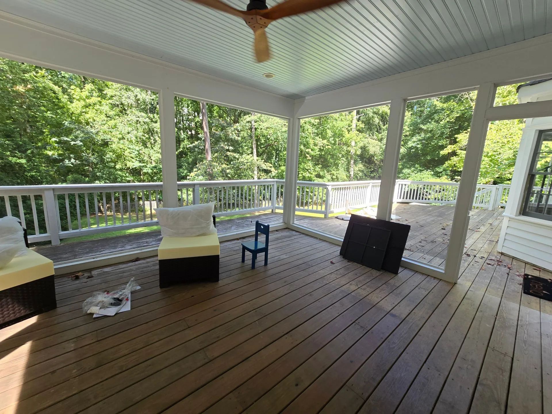 Covered porch with wooden floor, trees in the background, and a ceiling fan.
