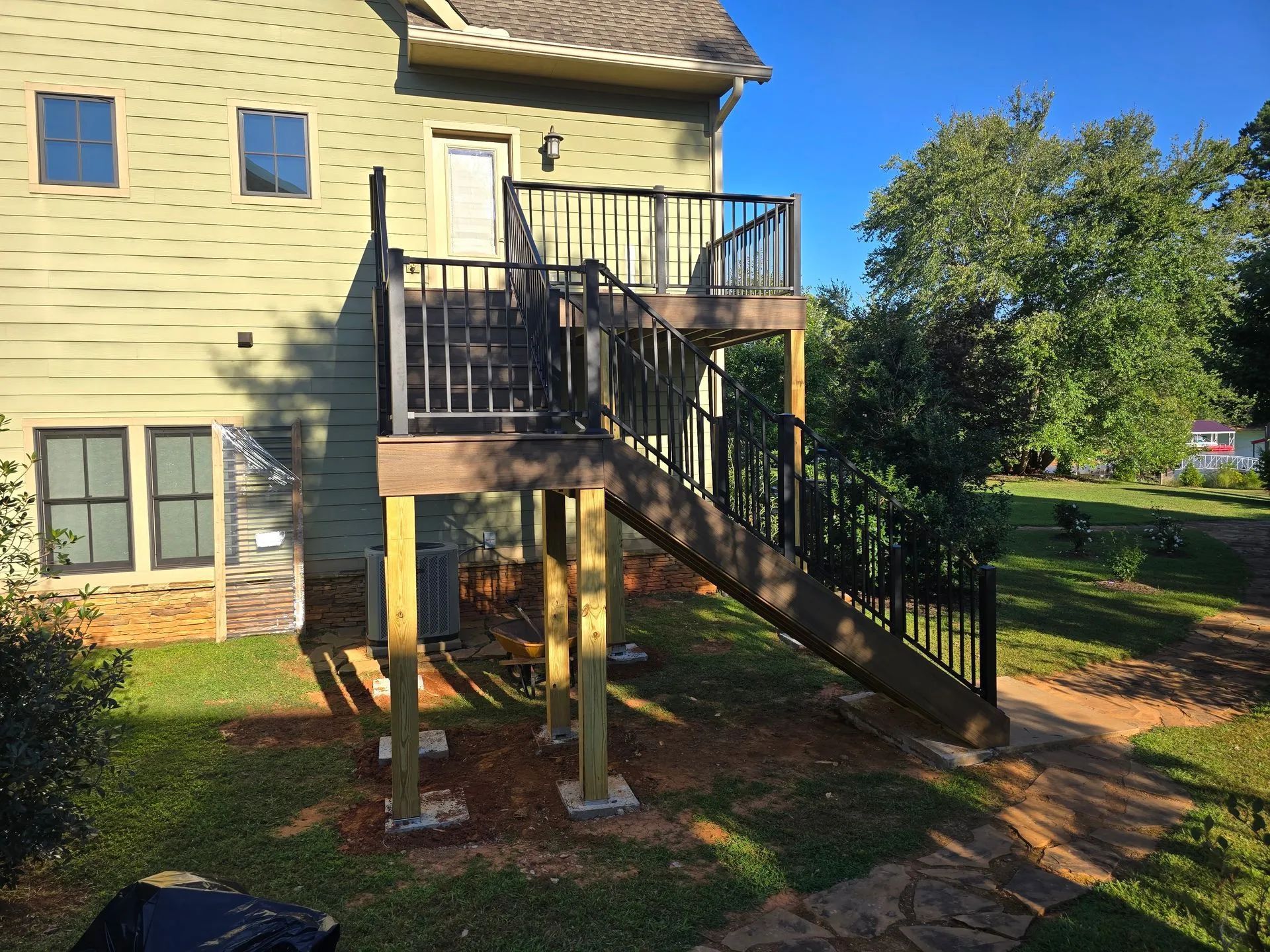 A two-level wooden deck with stairs, supported by posts, leading to a house with a light green exterior.