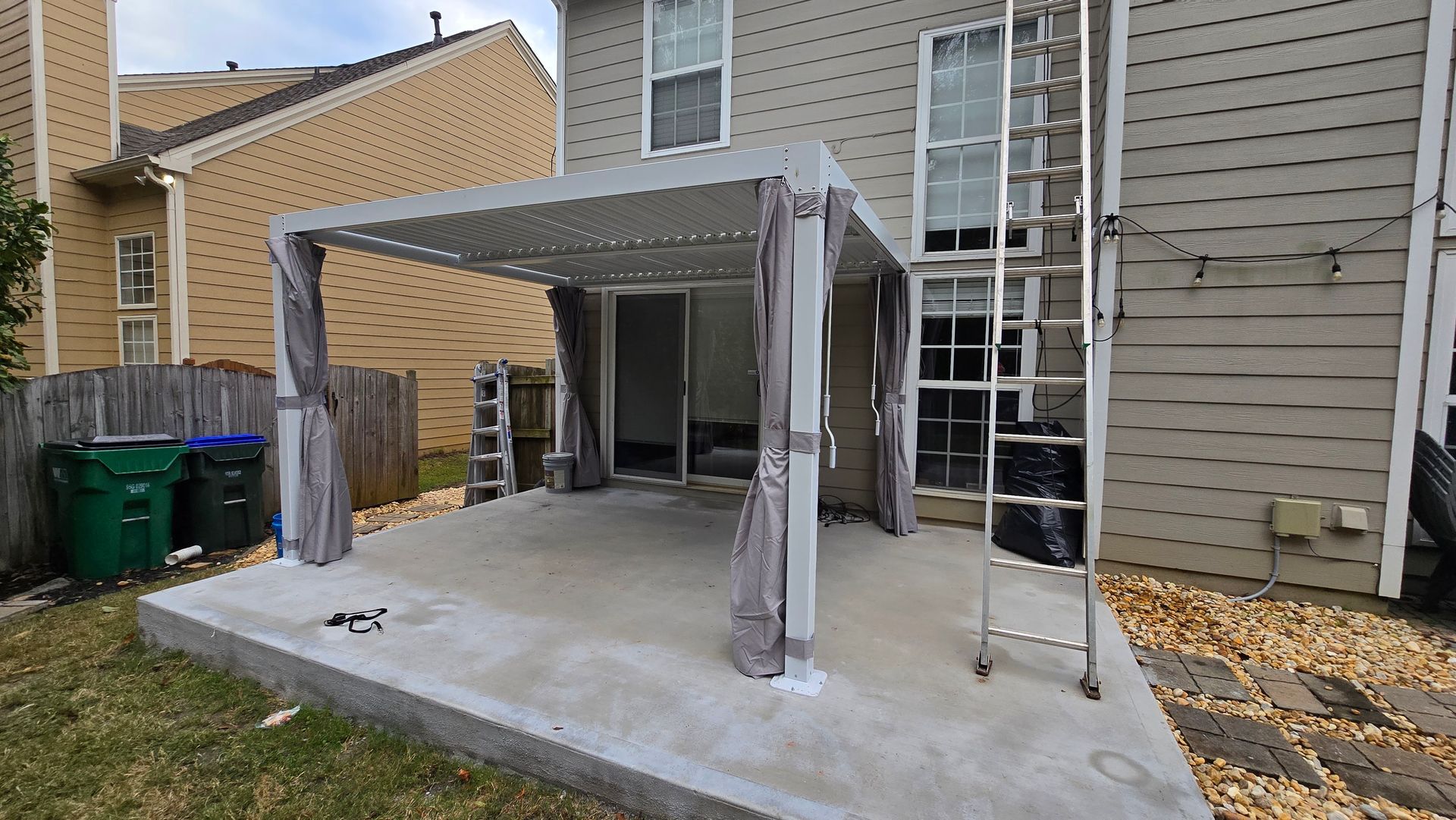 Patio with pergola and curtains, next to a house with a ladder leaning on it.