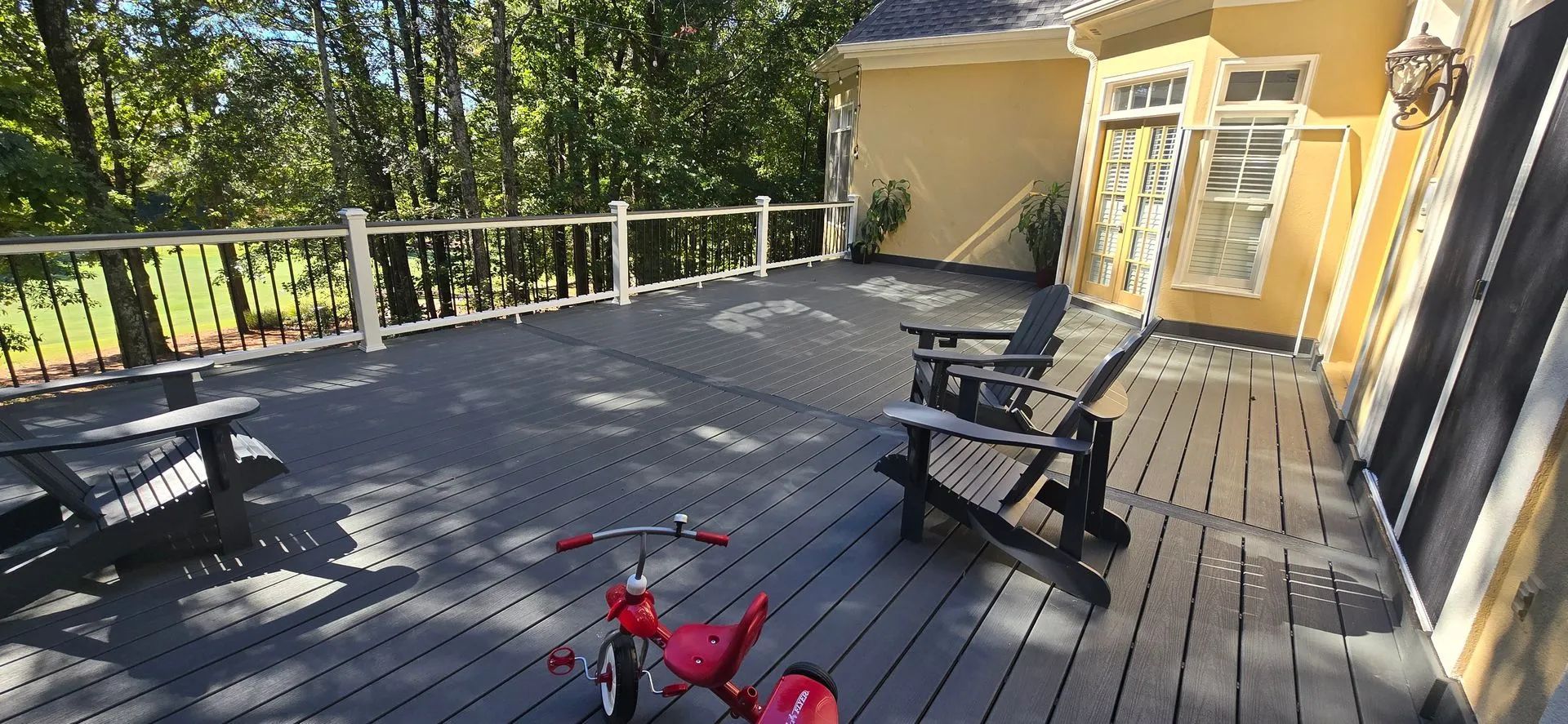 A red tricycle sits on a large wooden deck with black chairs and a picnic table.