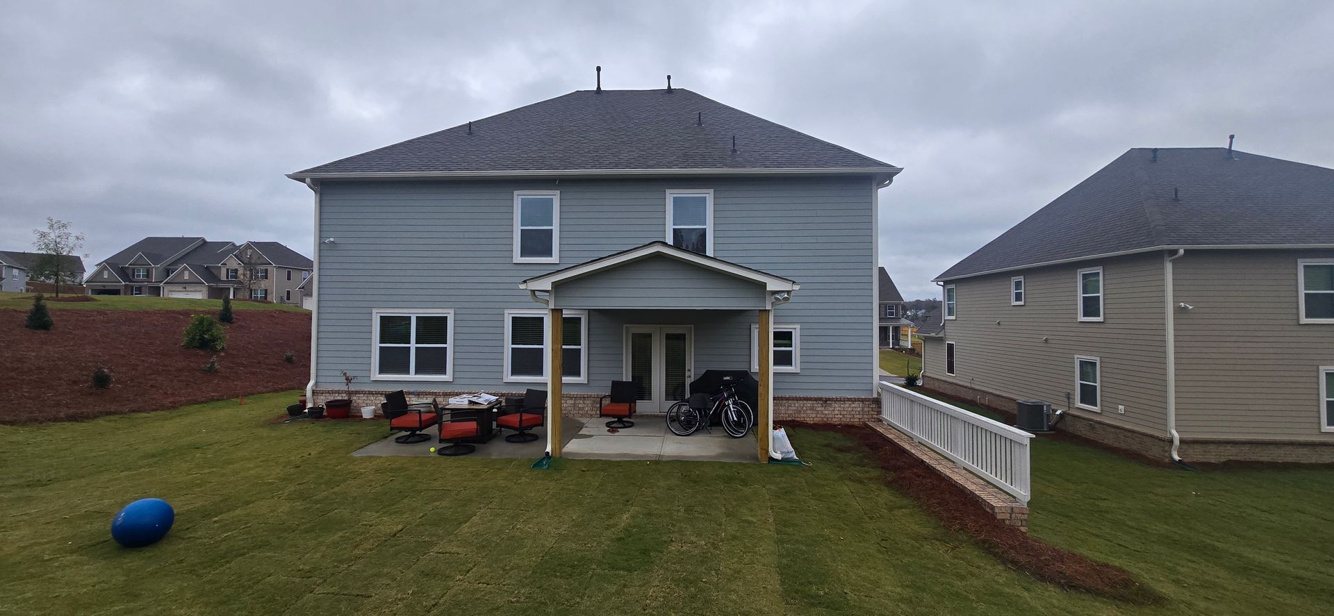 Back view of a two-story house with a patio, a blue ball in the yard, and a cloudy sky.