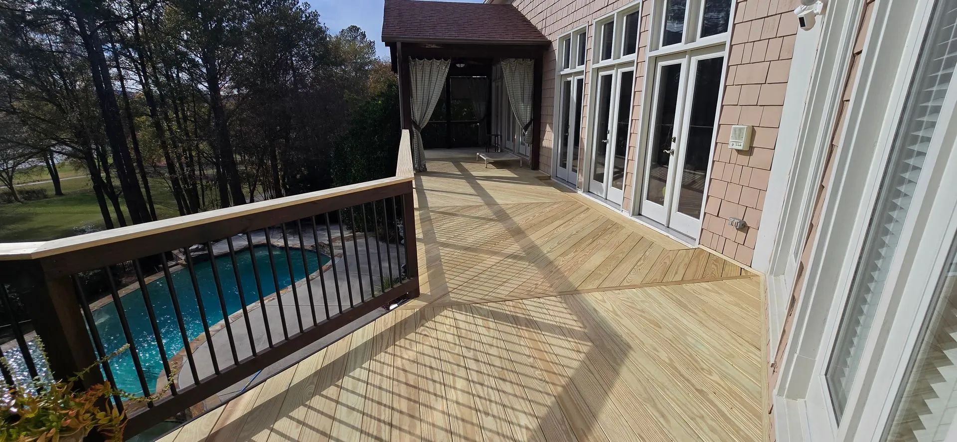 A wooden deck with a pool to the left. The deck leads to French doors on a house.