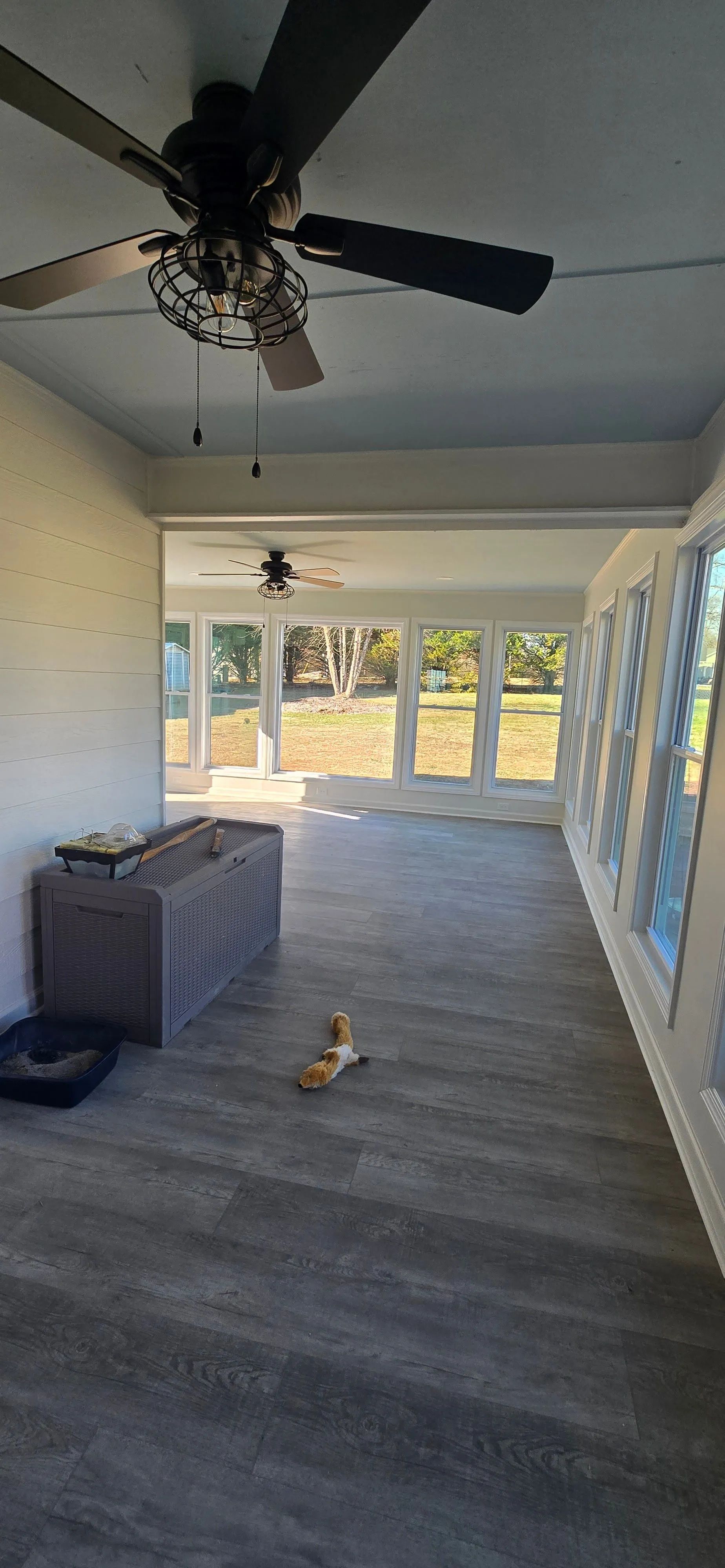 An enclosed sunroom with large windows, a ceiling fan, and a small dog on the floor.