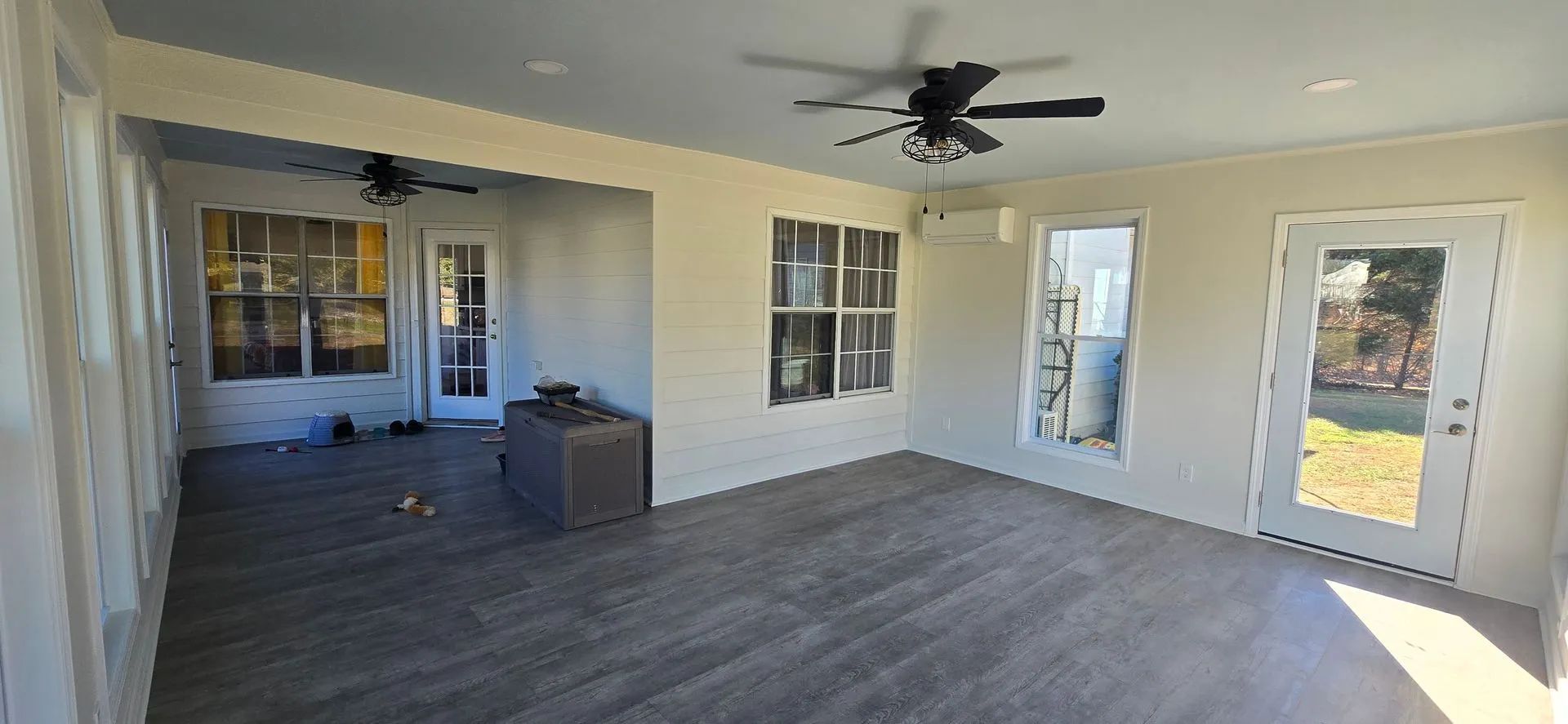 An enclosed porch with gray flooring, white walls, and a ceiling fan. There are windows and a door leading outside.