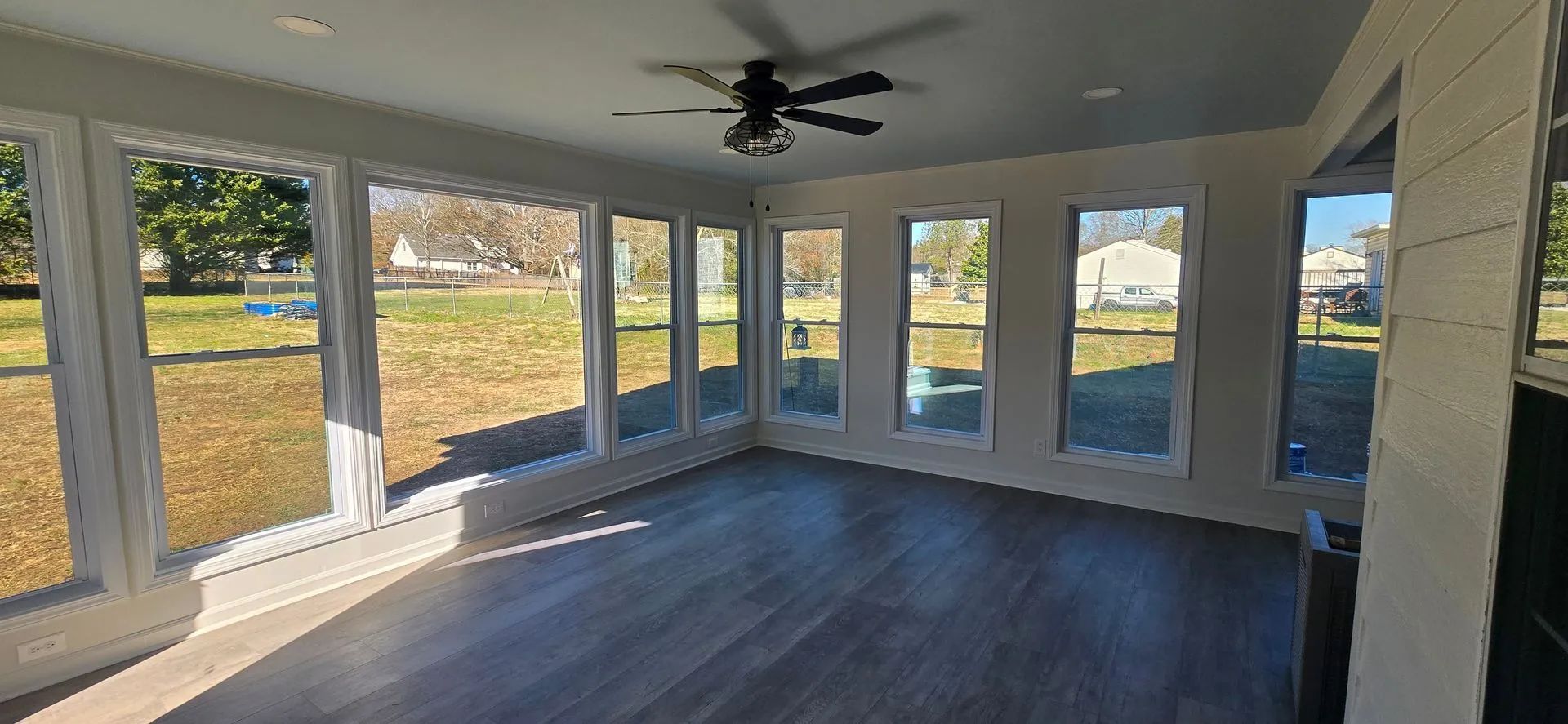 Sunroom with large windows overlooking a grassy yard. Gray flooring, white trim, and a ceiling fan.