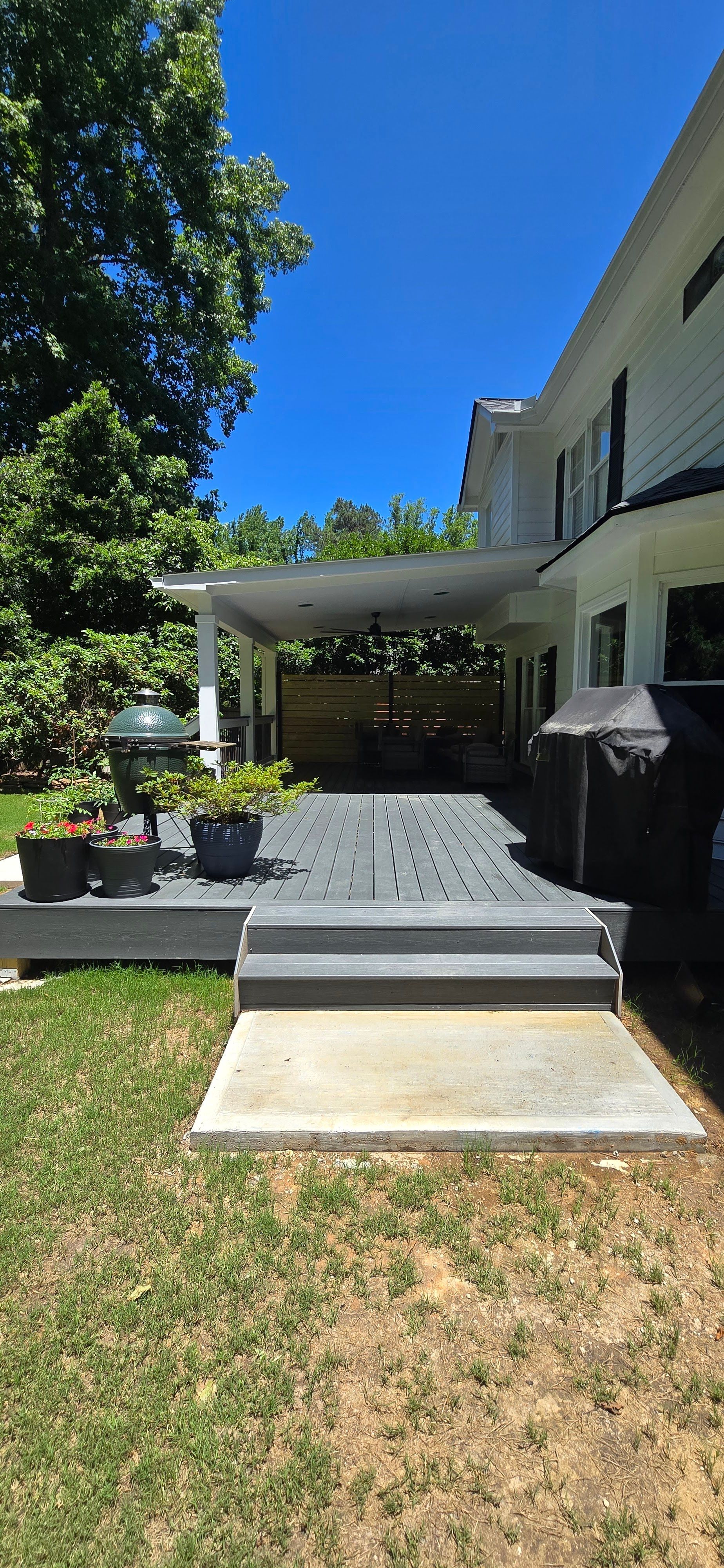 A backyard deck with steps leading to a covered patio, surrounded by green grass and trees.