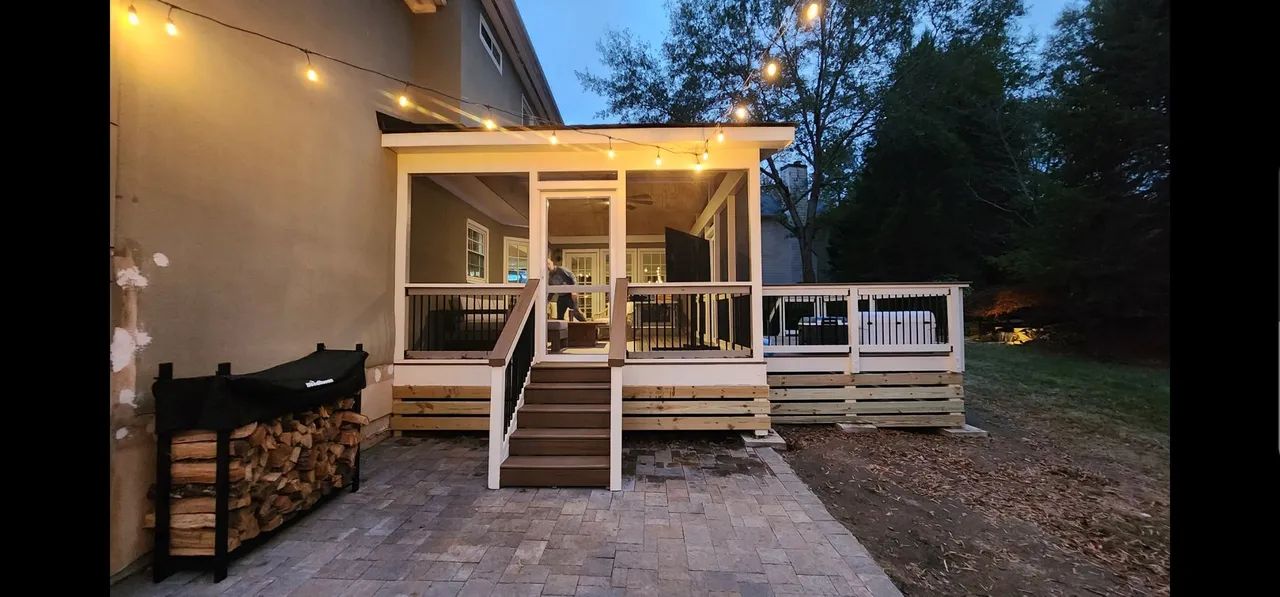 A screened porch with string lights, wooden deck, and firewood rack next to the house.