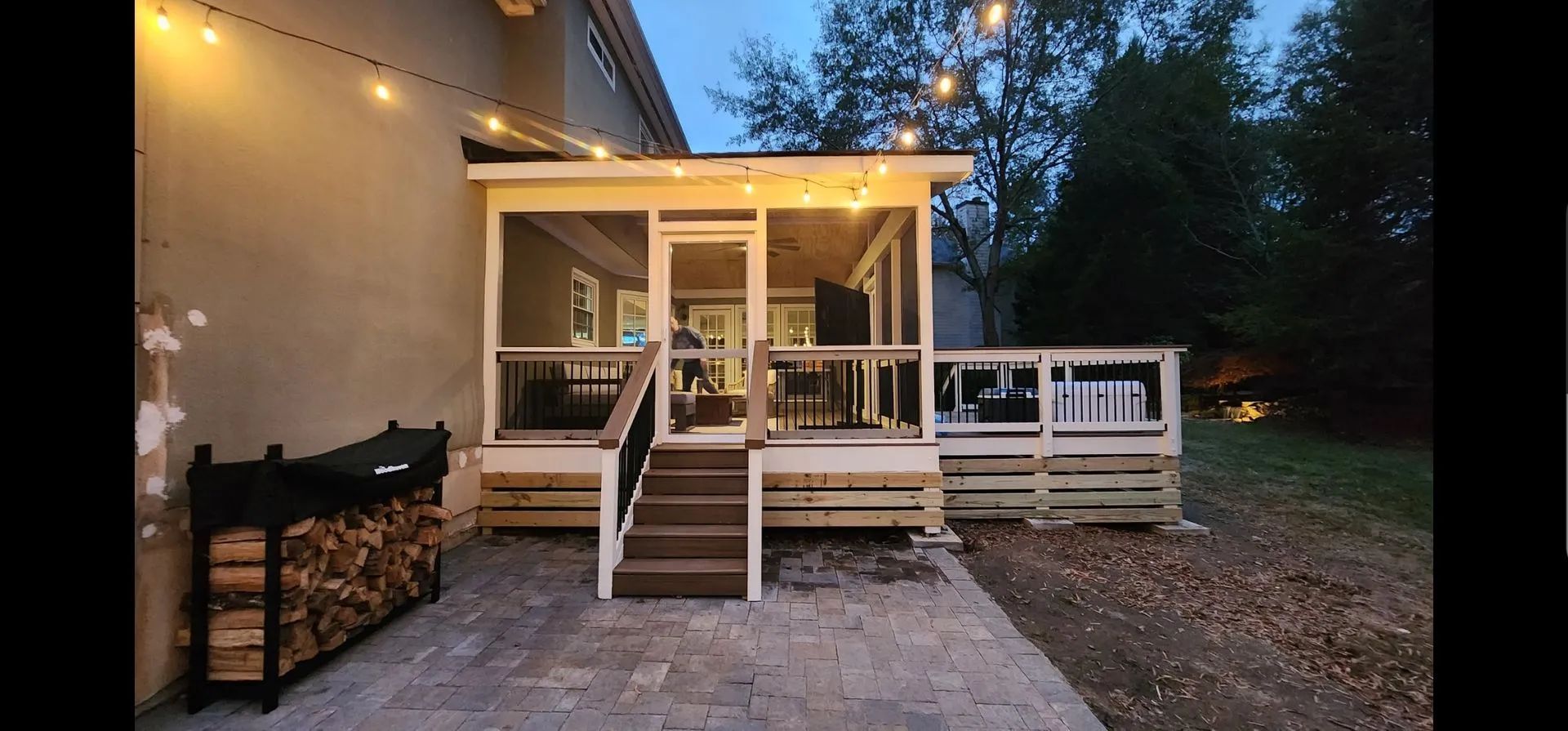 A screened porch with string lights, a deck, and a firewood rack, at dusk.