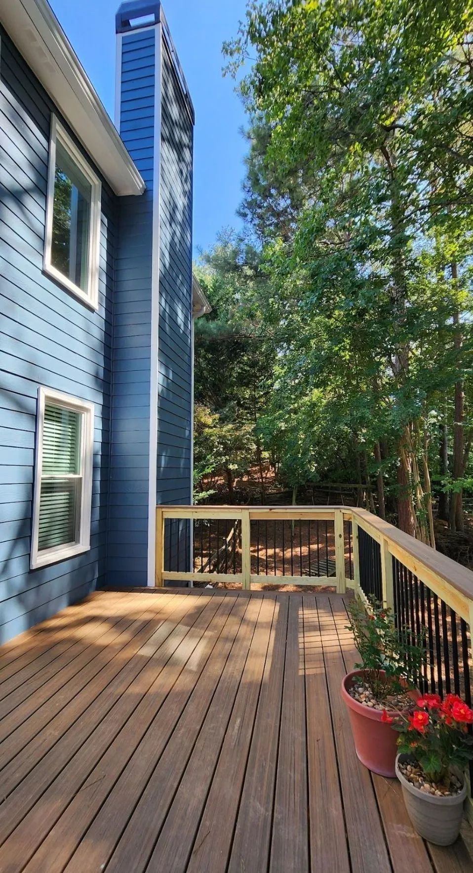 Wooden deck attached to blue house; black railing, trees in background.