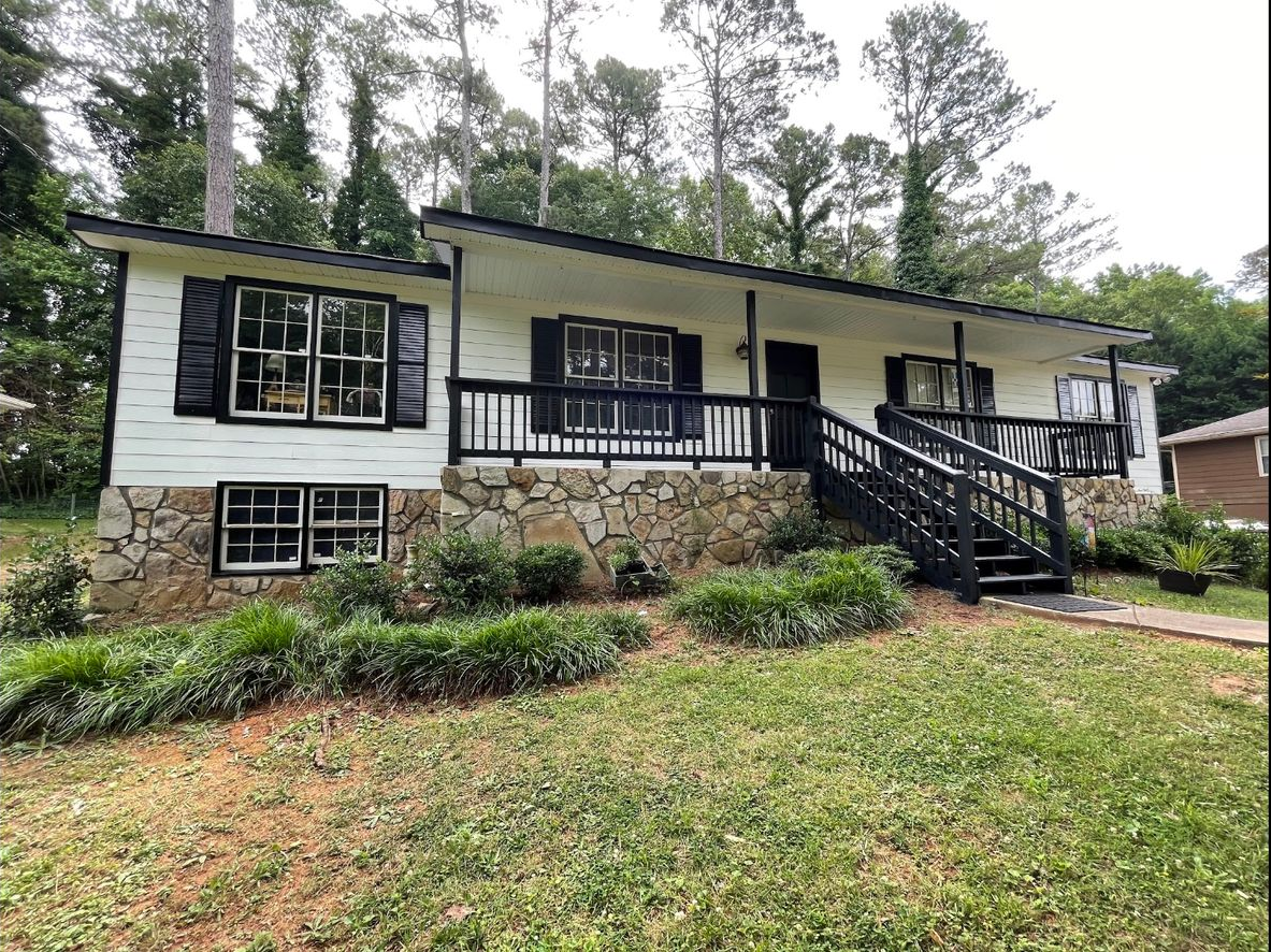 White house with black trim, stone foundation, and black stairs, surrounded by greenery.
