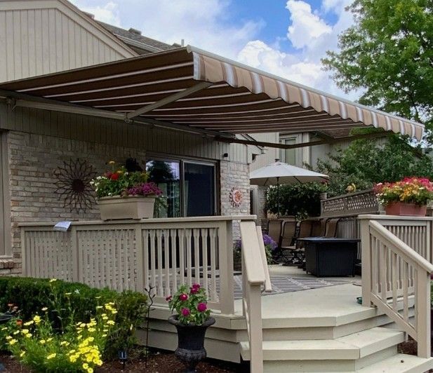 Black pergola with rolling shades, outside with trees and a white wall in the background.