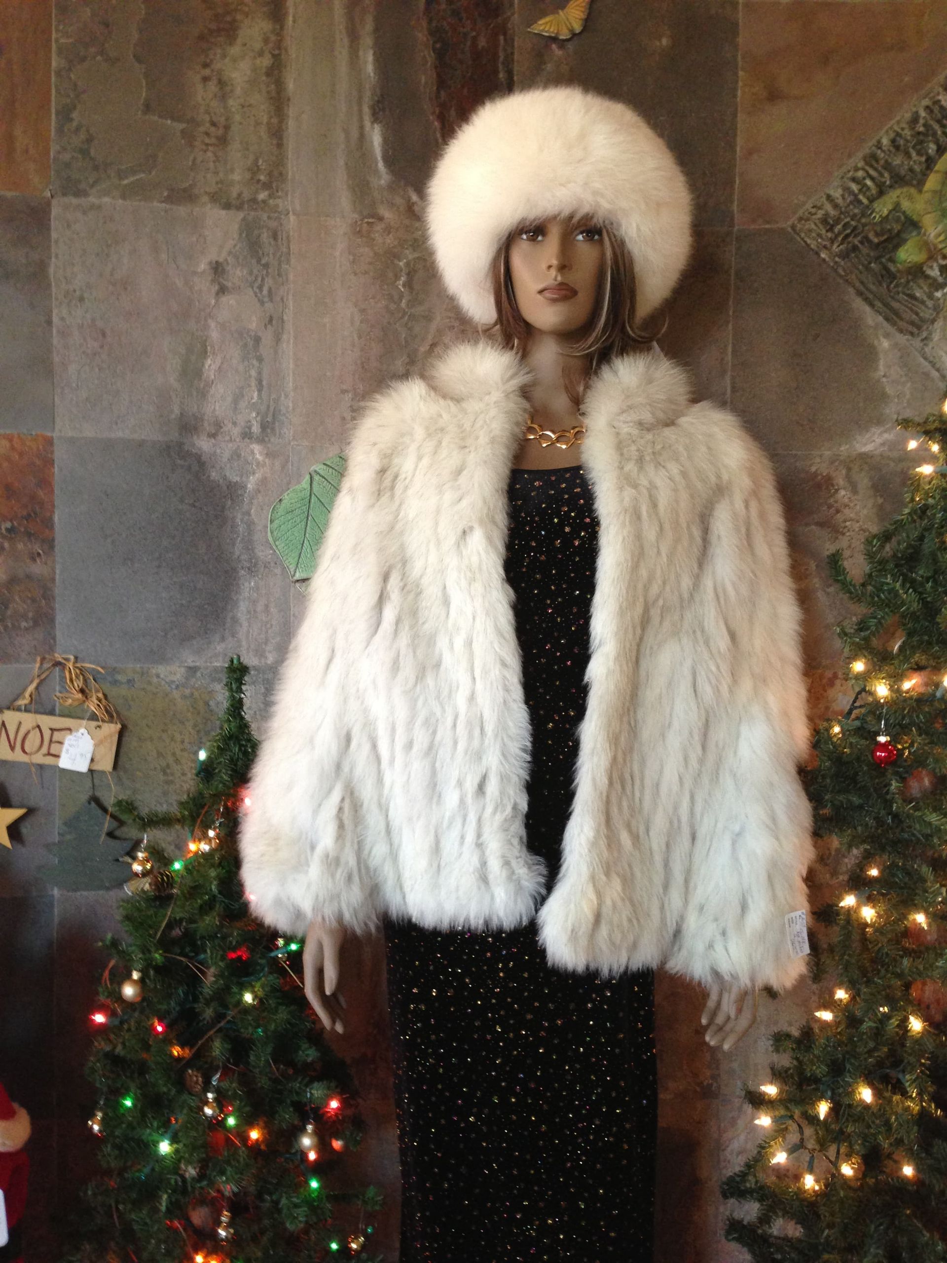 A mannequin wearing a white fur coat and hat is standing in front of a christmas tree.