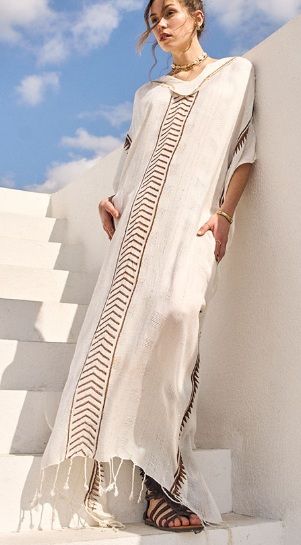 Mujer que lleva un vestido largo blanco con ribetes estampados de color marrón, apoyada contra una pared blanca.