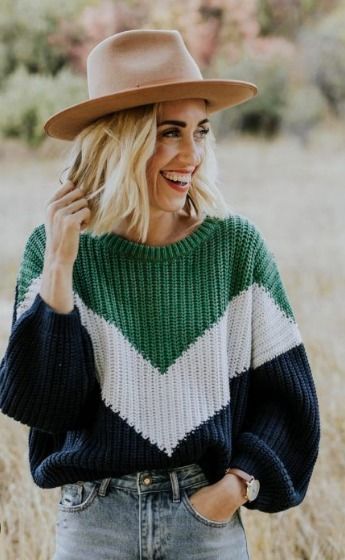 Una mujer con un suéter verde, blanco y azul marino y un sombrero marrón sonríe al aire libre.