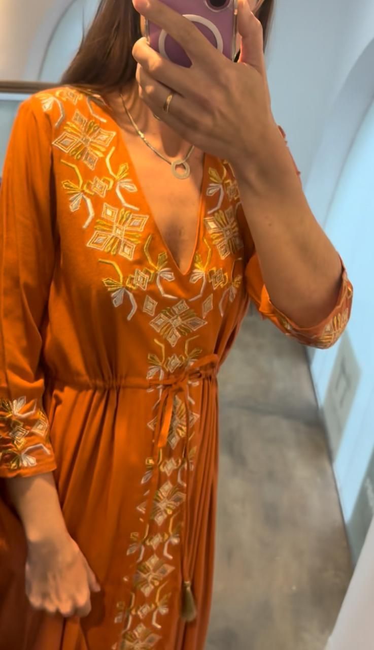 Mujer con un vestido largo naranja cruzado con bordados dorados, parada frente a un espejo.