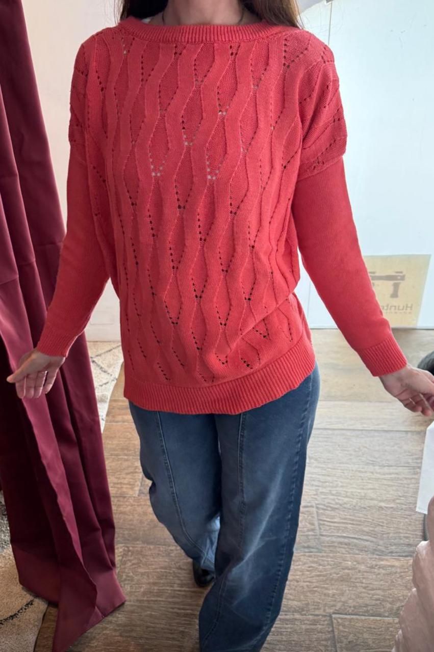 Mujer con suéter coral y jeans azules. De pie en un espacio interior.