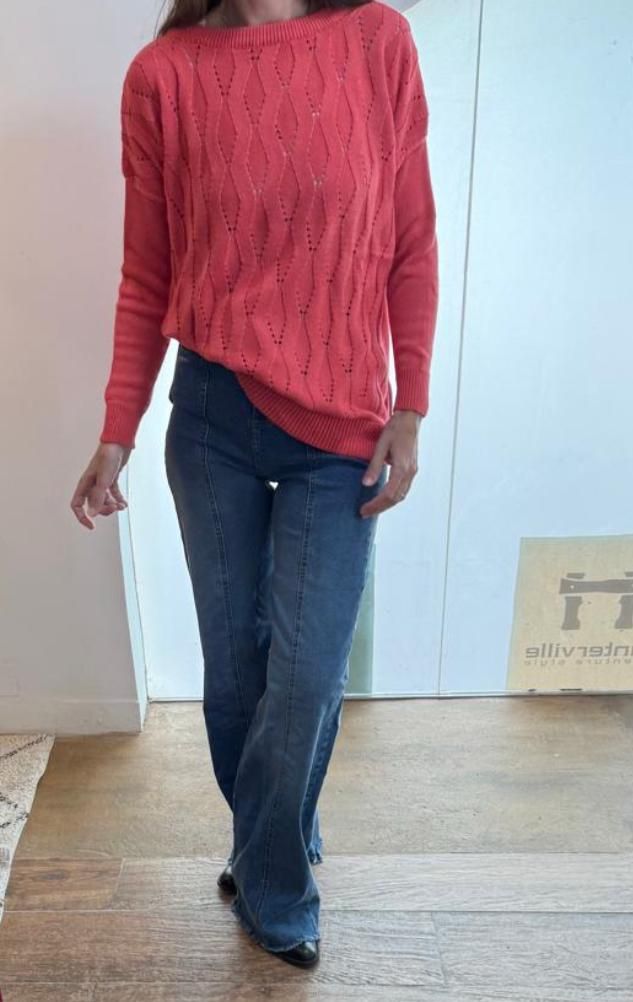 Mujer con suéter coral y jeans azules. De pie en un espacio interior.