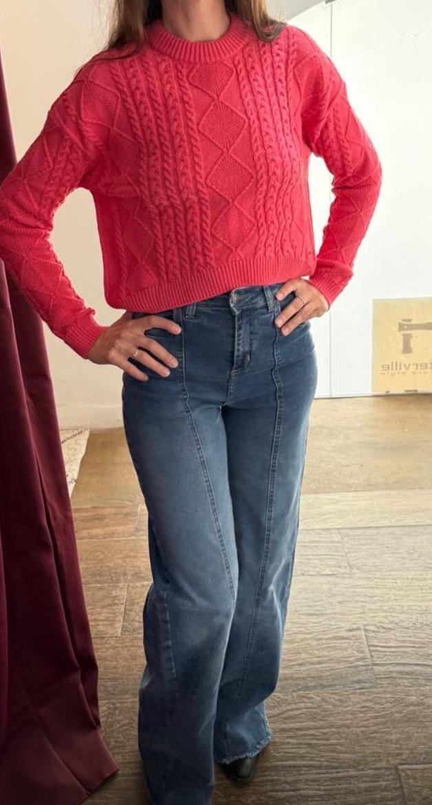 Mujer con suéter rojo y jeans azules posa con las manos en las caderas.