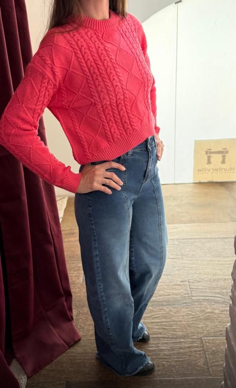 Mujer con suéter rojo y jeans azules posa con las manos en las caderas.