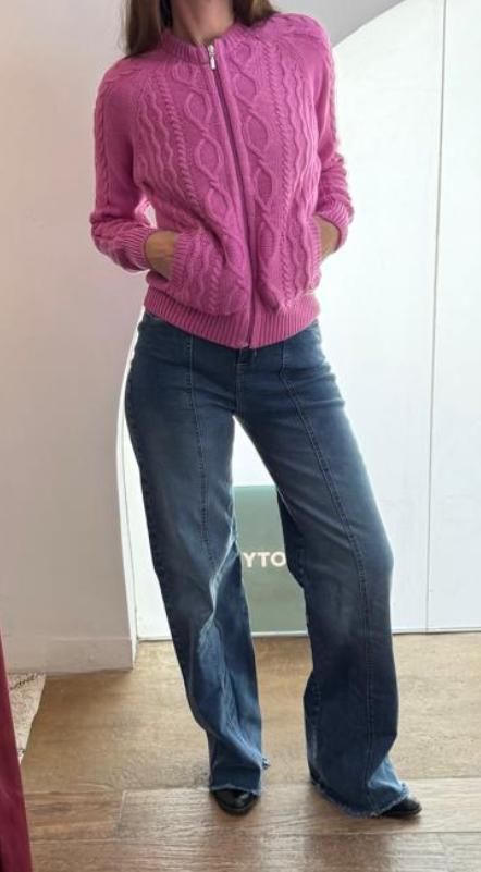 Mujer con suéter rosa con cremallera y jeans azules, parada en una habitación, con las manos en los bolsillos.