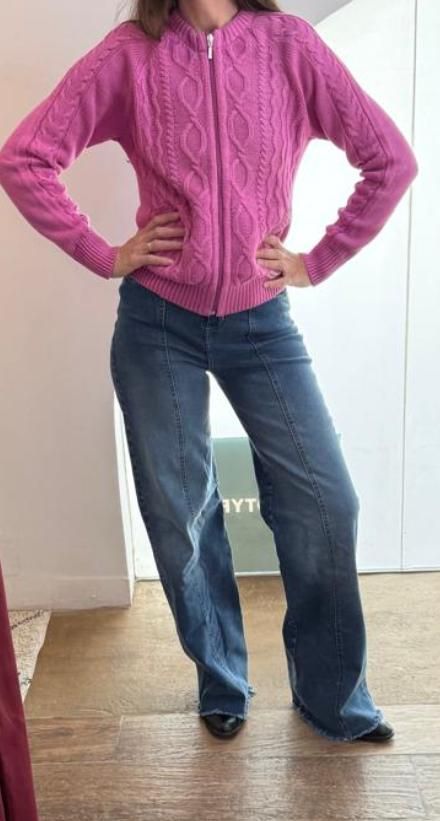 Mujer con suéter rosa con cremallera y jeans azules, parada en una habitación, con las manos en los bolsillos.