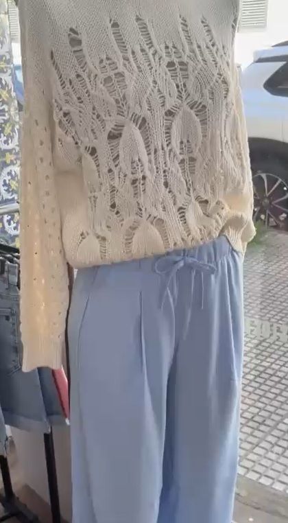 Suéter de punto abierto, de color crema y cuello redondo, para usar sobre un pantalón azul claro con cintura anudada.