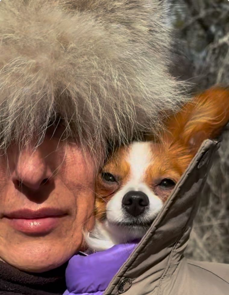 Person in a furry hat holding a small white-and-brown dog in a purple jacket outdoors