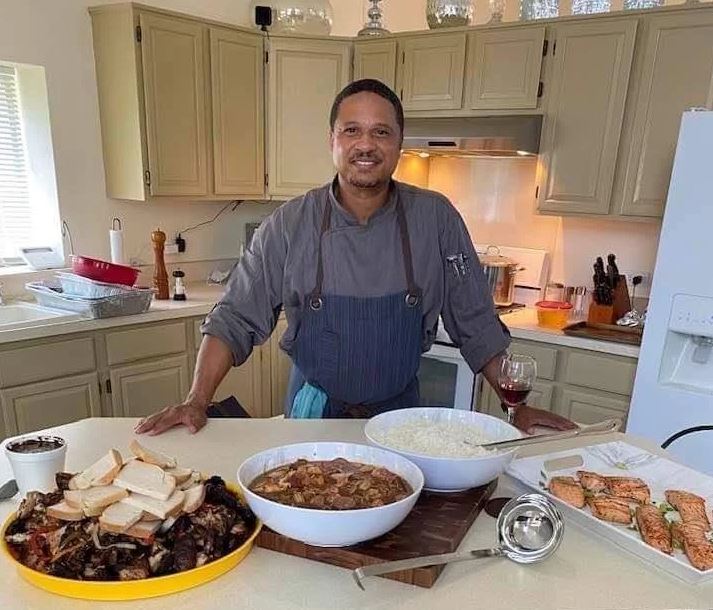 A man in an apron is standing in a kitchen with bowls of food on the counter.