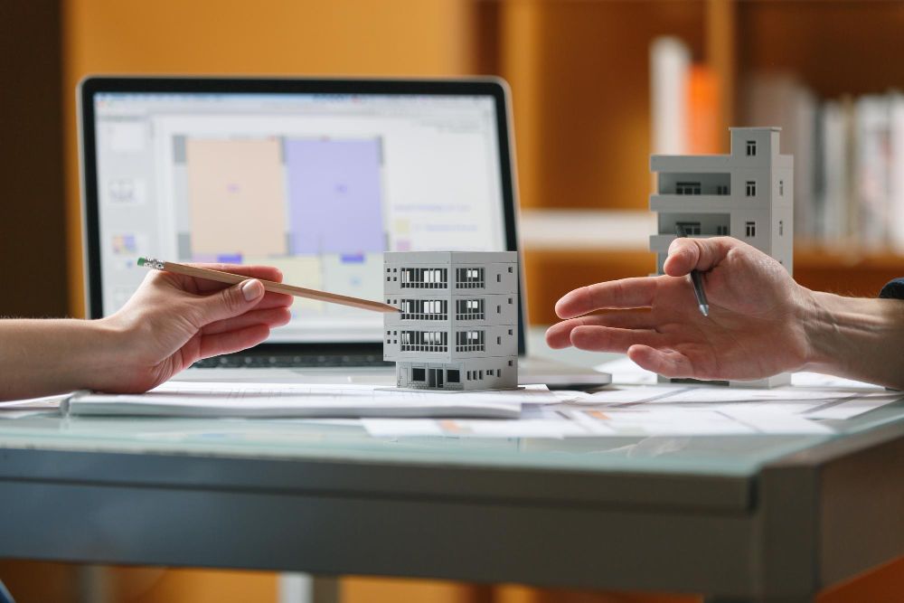 Two people are sitting at a table with a laptop and a model house.