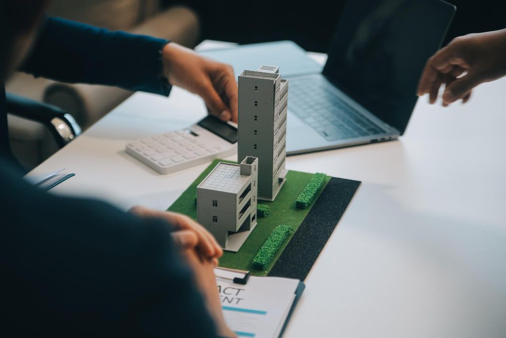 Two people are sitting at a table with a model house and a clipboard.