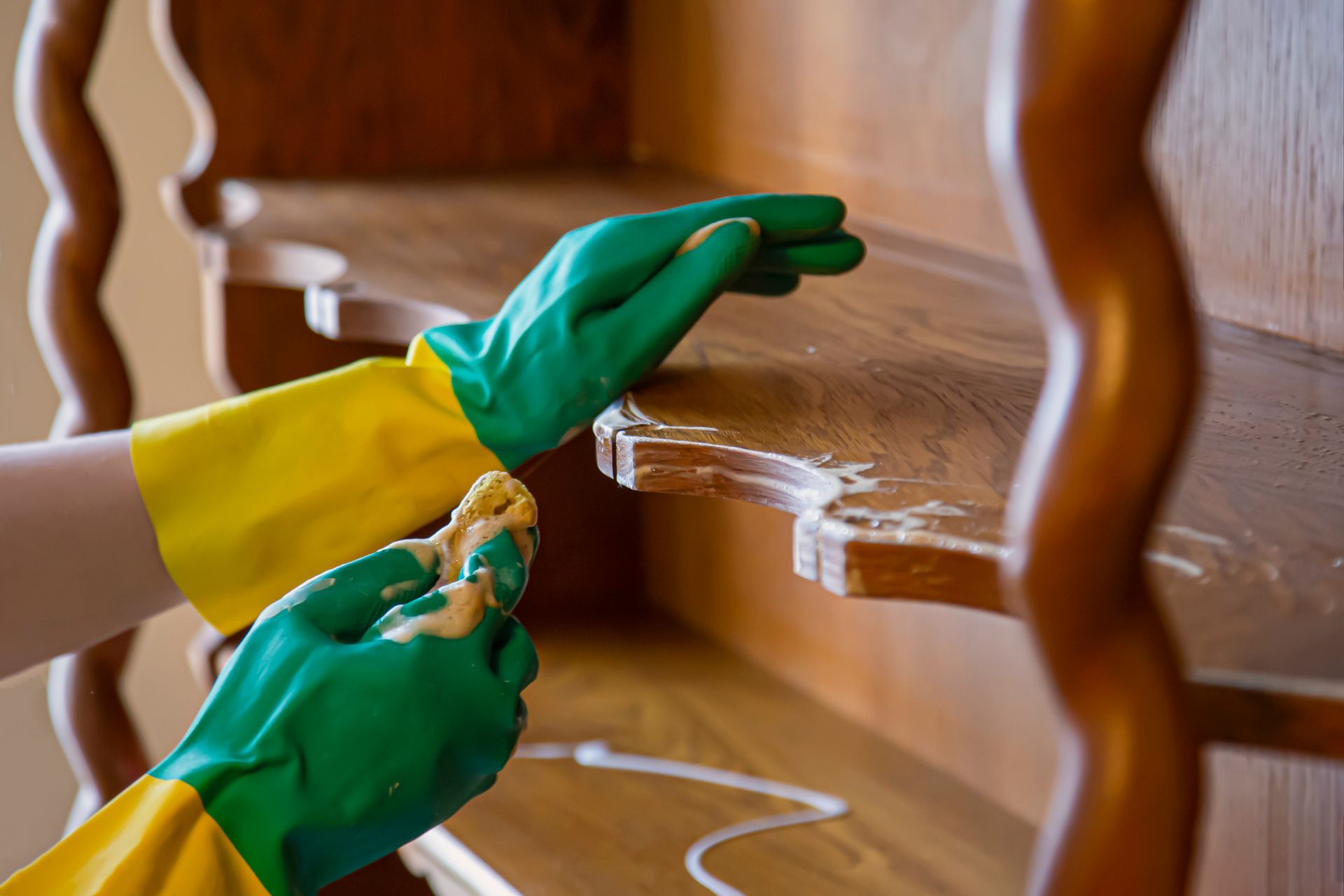 A person wearing green and yellow rubber gloves is cleaning a wooden shelf.